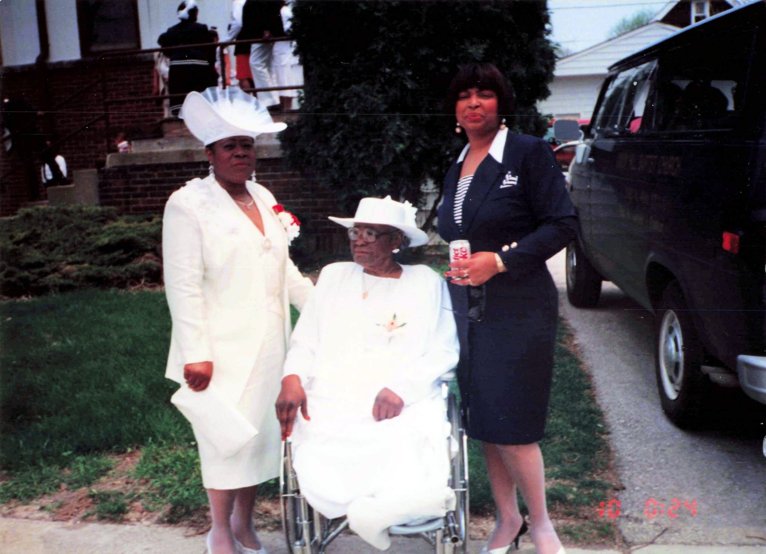 Three women in formal outfits outdoors, one elderly woman seated in a wheelchair, two others standing beside her near a parked vehicle.