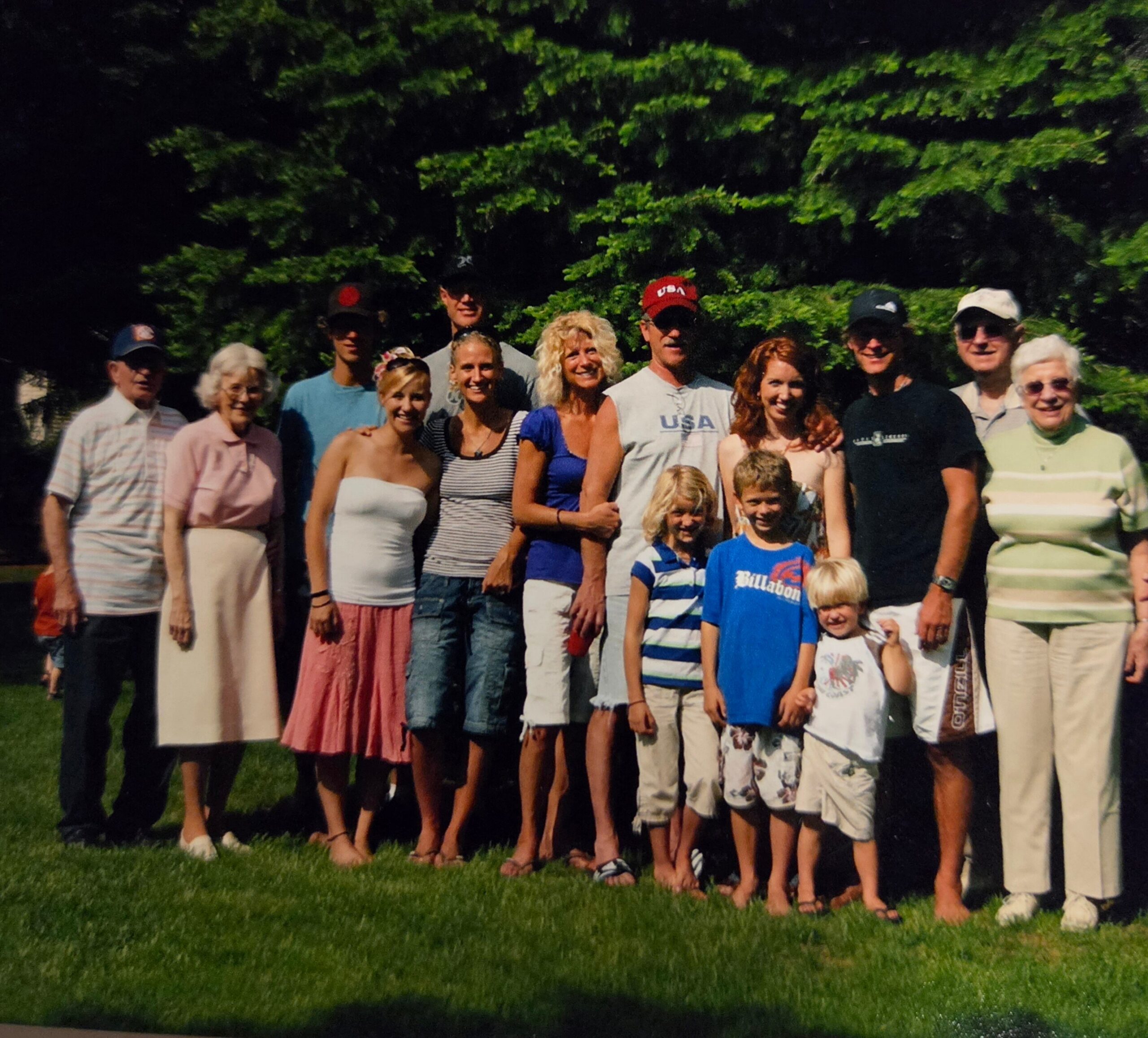 Group photo of adults and children standing together on a grassy lawn with trees behind them, smiling for the camera at an outdoor gathering.