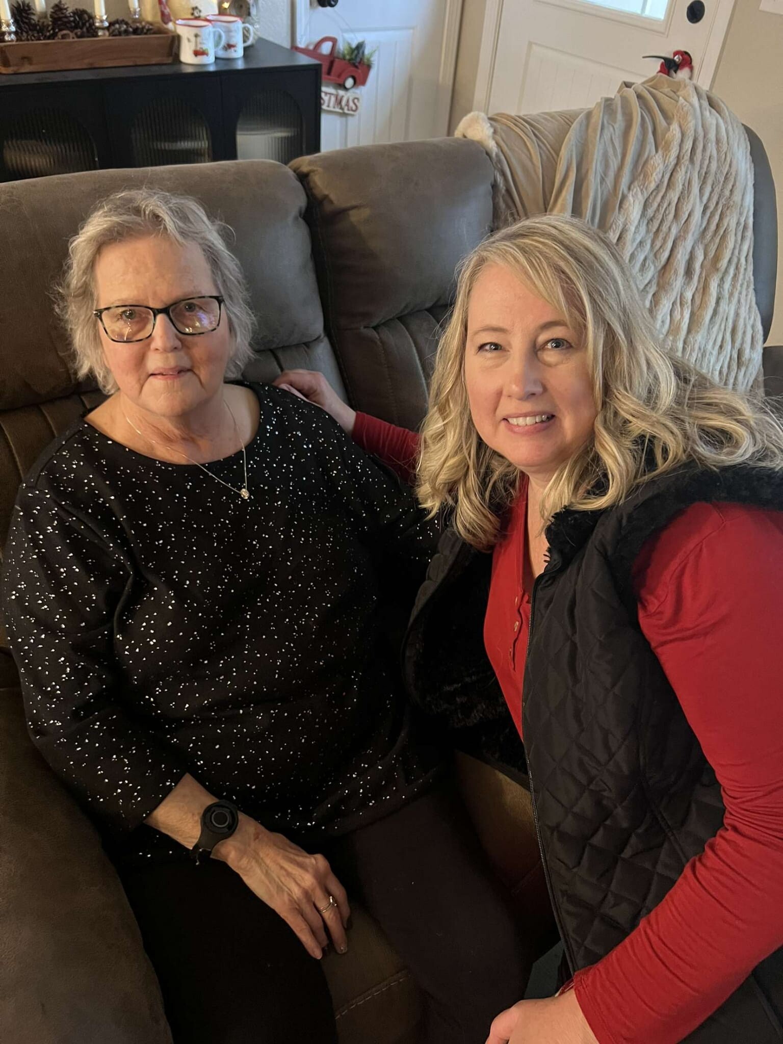 Two women sit close together on a brown couch in a cozy living room, one with glasses wearing a black speckled top, the other in red with a black quilted vest, smiling at the camera.
