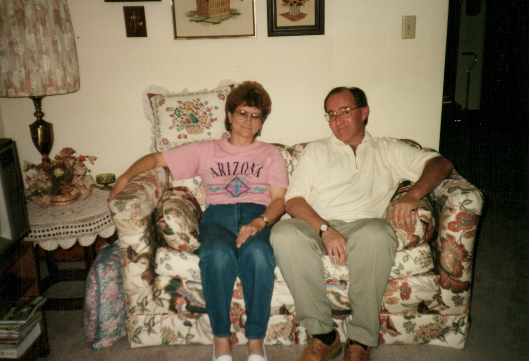 Two adults sit together on a floral-patterned couch in a cozy living room, relaxed and smiling.