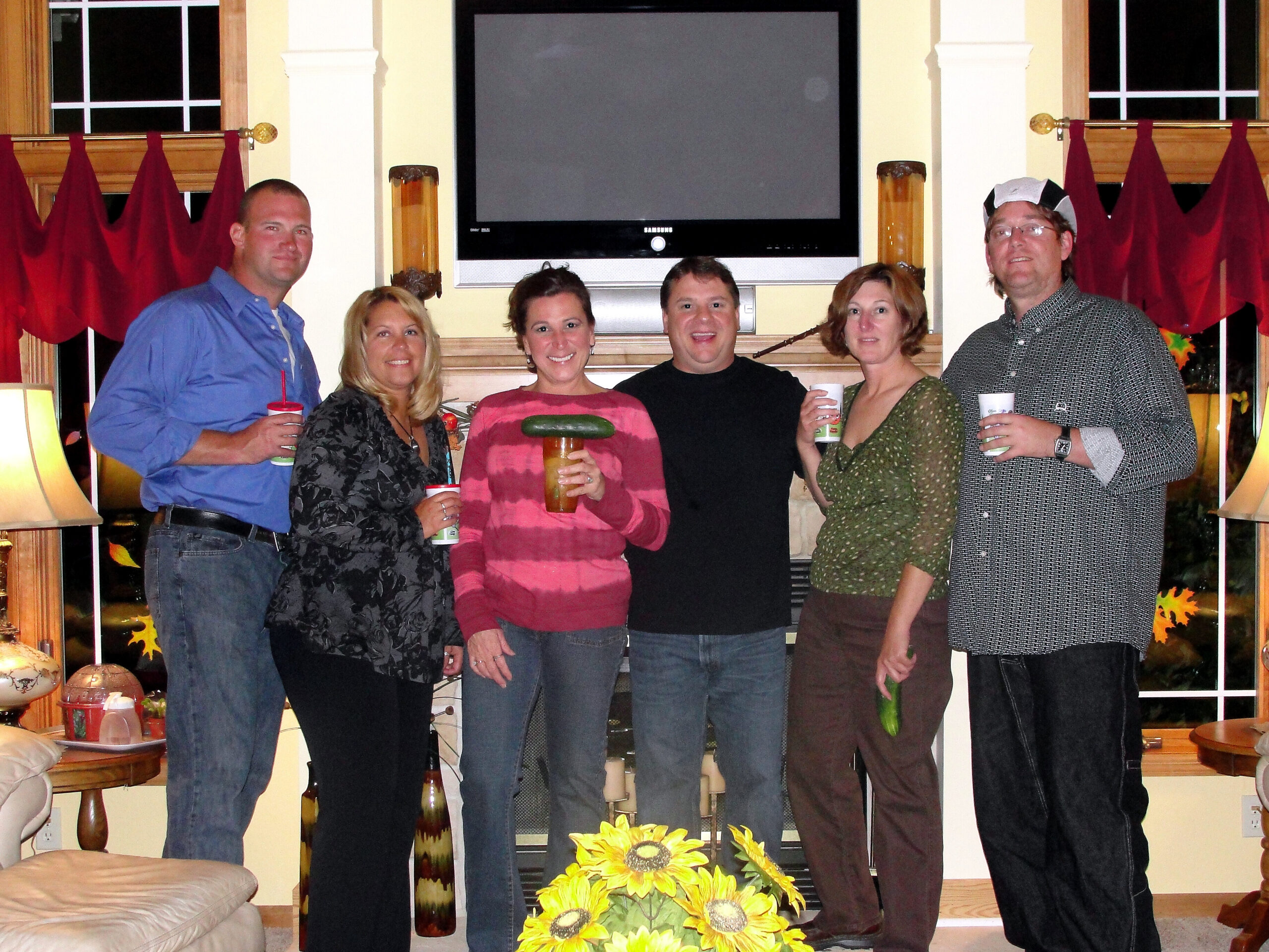 Six adults posing for a group photo in a living room, holding drinks, with a large TV behind them and sunflowers in the foreground.
