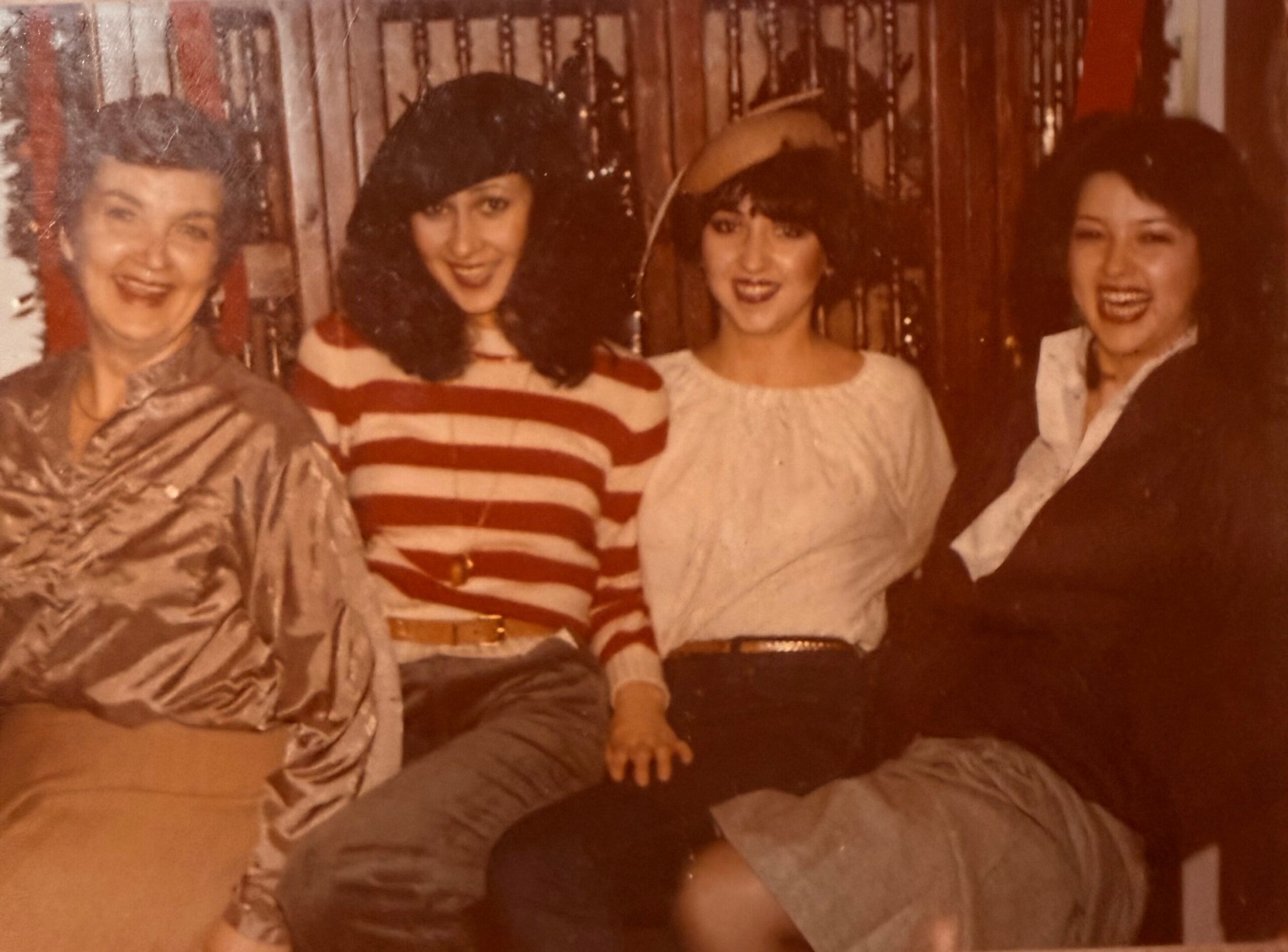 Four women pose together at a social gathering, smiling at the camera; left to right: metallic blouse, red-and-white striped sweater, white top, dark blazer.