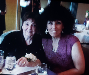 Two women sit at a restaurant table, smiling at the camera; left in black with a white corsage, right in a purple lace dress.