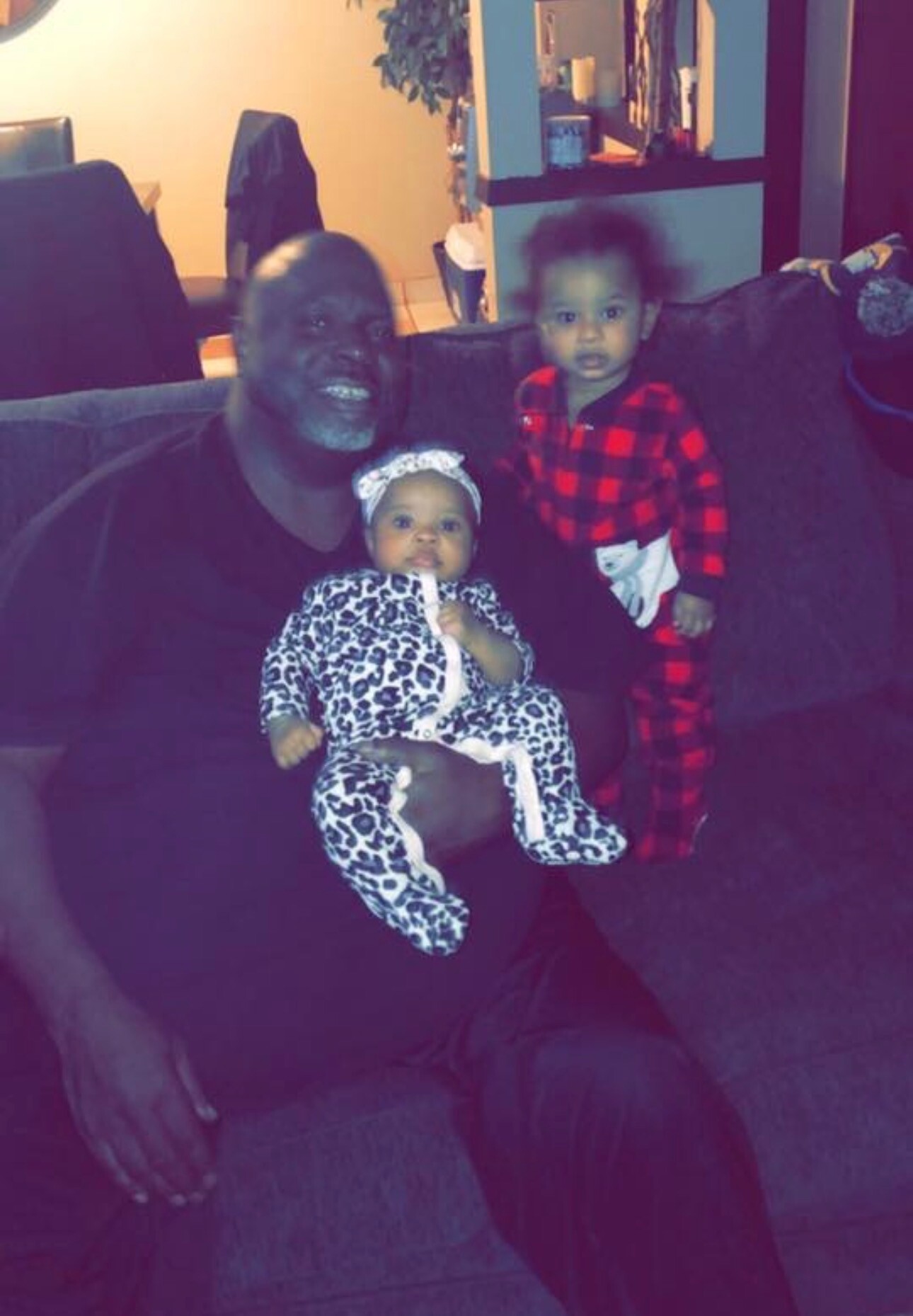 A Black man sits on a sofa holding a baby in a leopard-print onesie while a toddler in red plaid pajamas stands beside them in a living room.