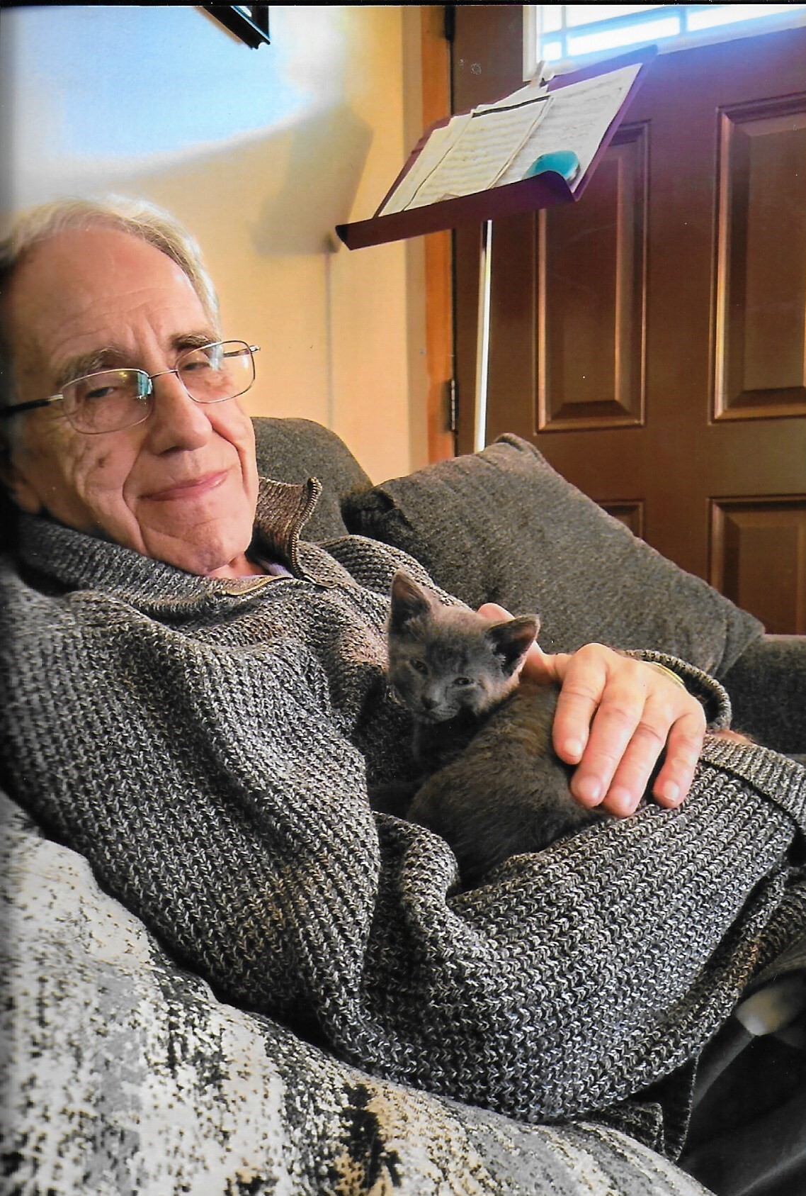 Elderly man wearing a gray sweater sits on a sofa, smiling, while holding a small gray kitten on his lap.