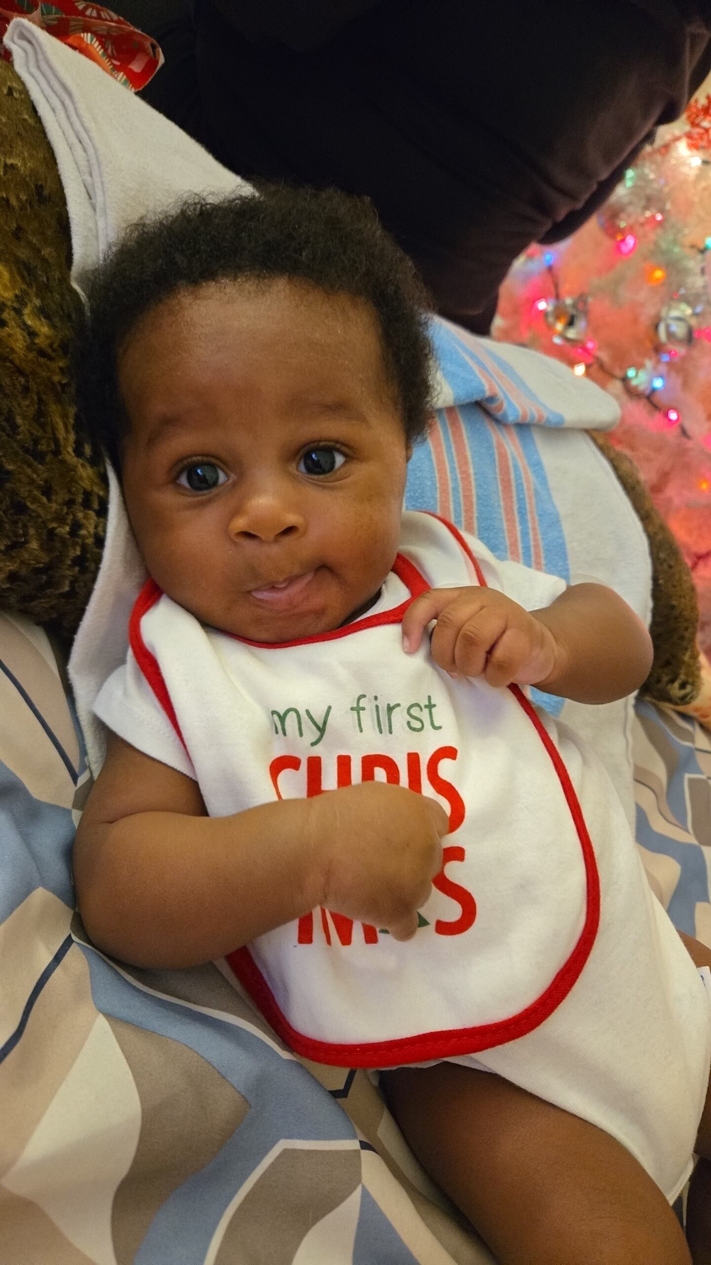 Close-up of a baby with big eyes wearing a white bib with red trim that reads 'my first Christmas,' held by a caregiver with a patterned blanket, a pink Christmas tree with colorful lights in the background.