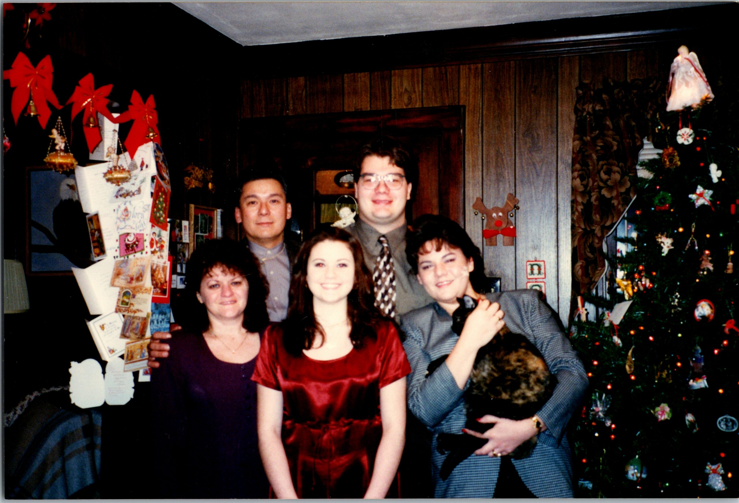 Group of five adults posing for a Christmas photo indoors, with a decorated tree and holiday cards on the left.