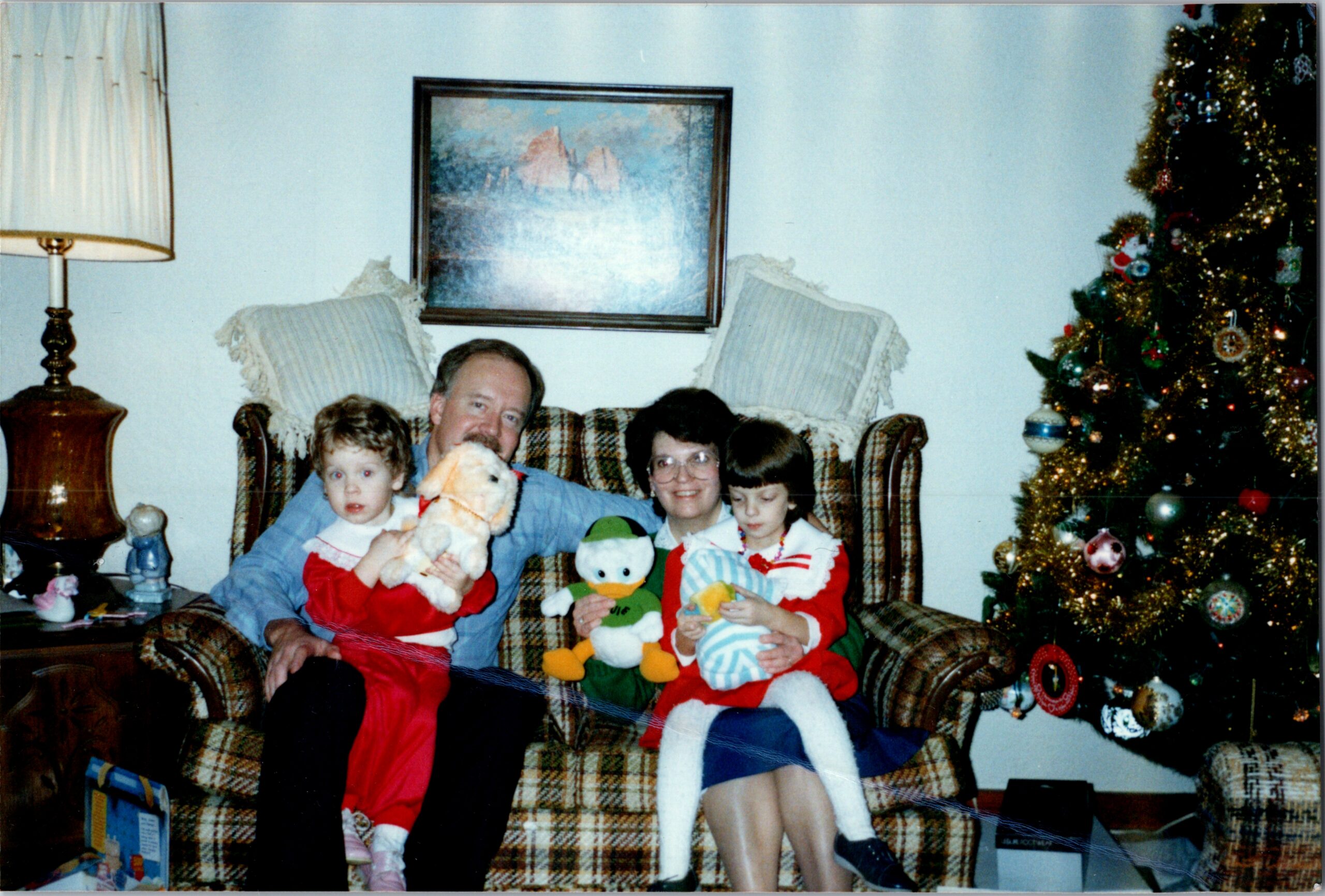 Family sits on a plaid sofa with kids, holding stuffed toys, Christmas tree visible on the right in a cozy living room.