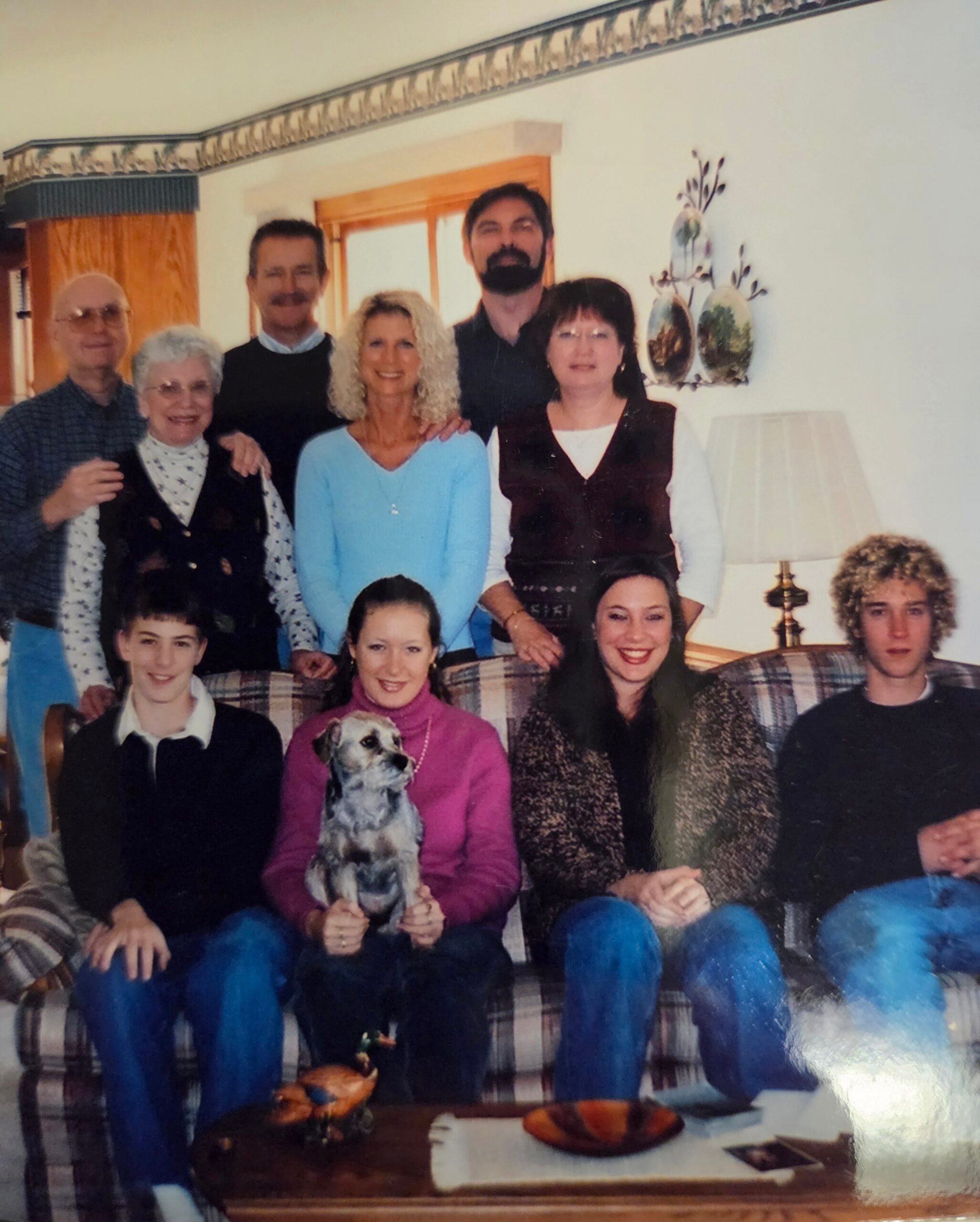 Family group posing on a plaid sofa in a living room; adults stand behind several seated younger people, with a dog sitting in front center.