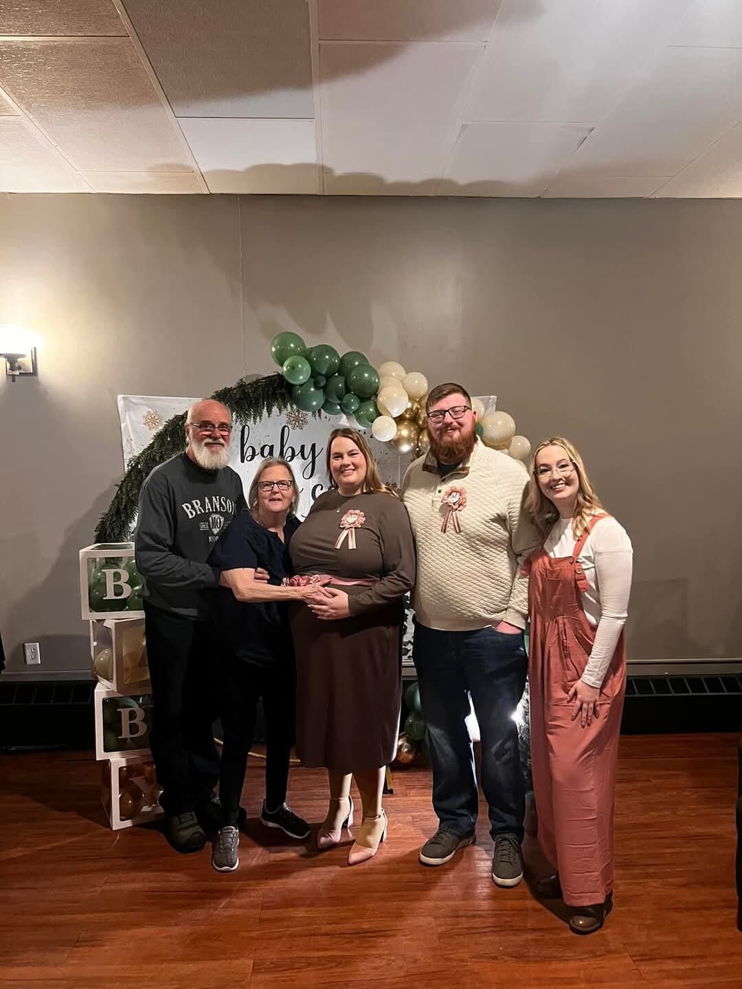 Group of five amigos posing for a baby shower photo with green, white, and gold balloon arch and a ‘baby’ sign in the background; pregnant woman center wearing a brown dress and ribbon.