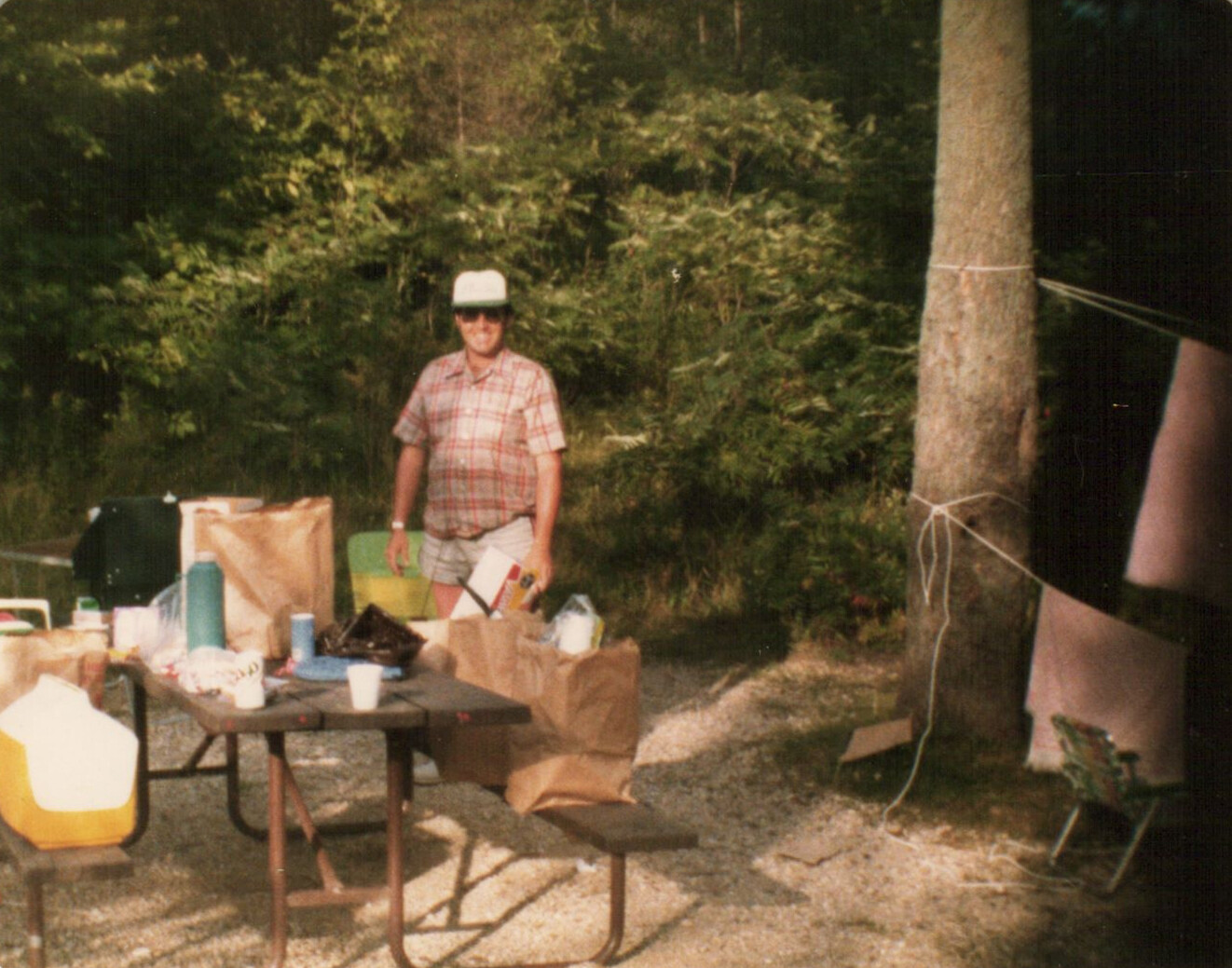 Man in a plaid shirt and white cap standing beside a table piled with bags and camping supplies, trees in the background.