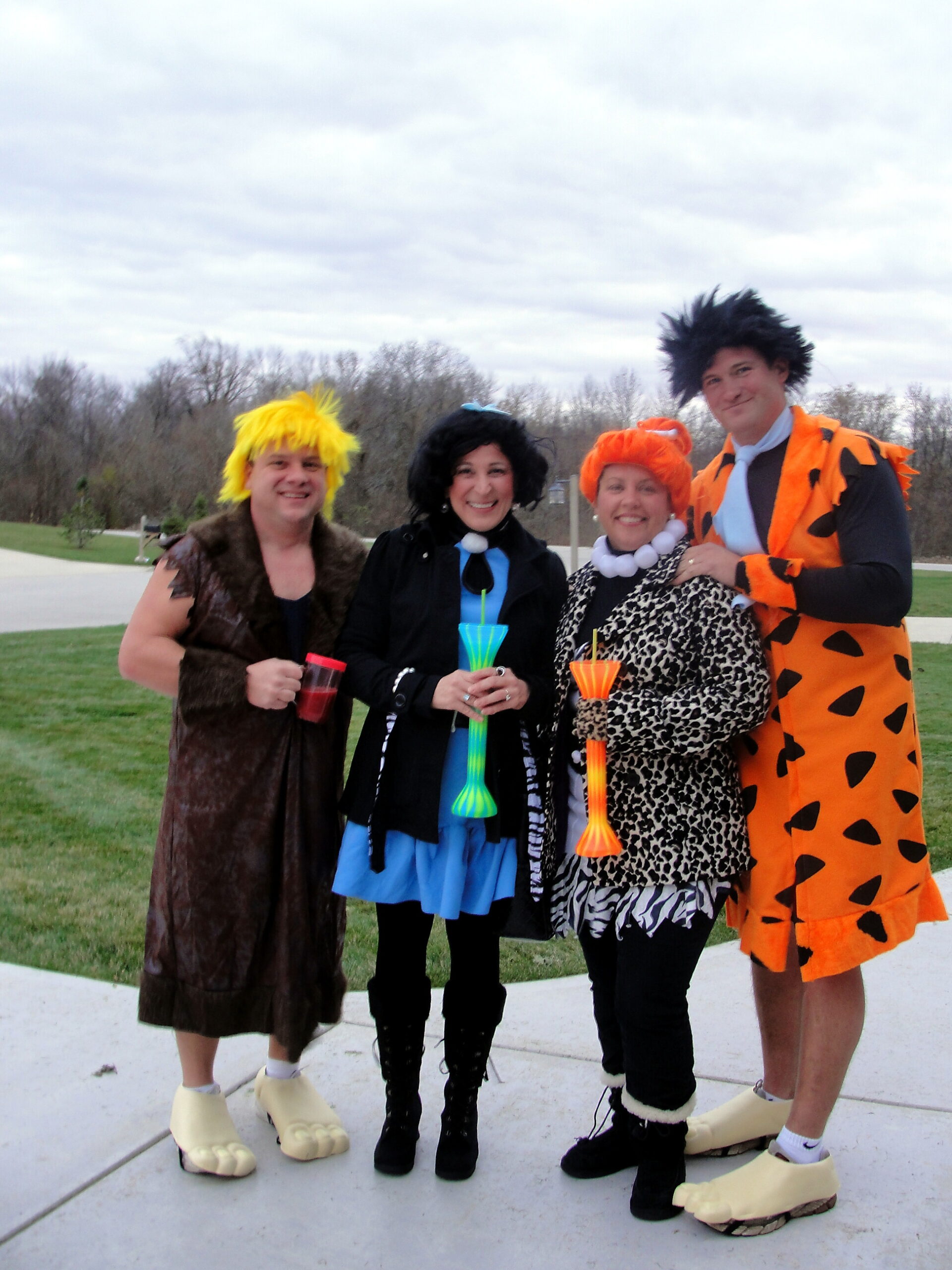Four adults in silly costumes pose outdoors, each holding colorful drinks and wearing oversized foam feet.