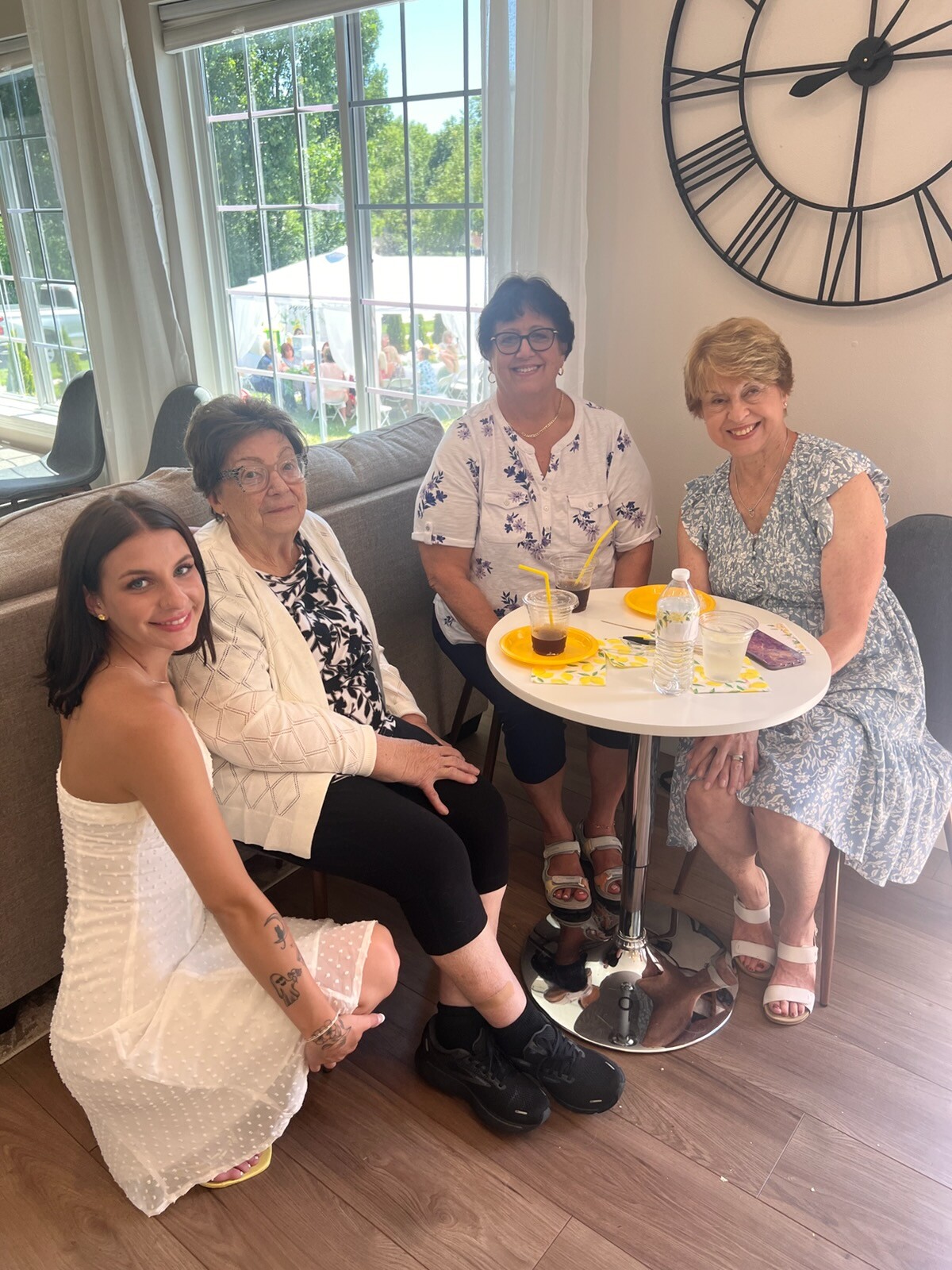 Four women sit around a small round table indoors, smiling with drinks and yellow plates, large wall clock behind them and a sunny outdoor scene visible through windows.