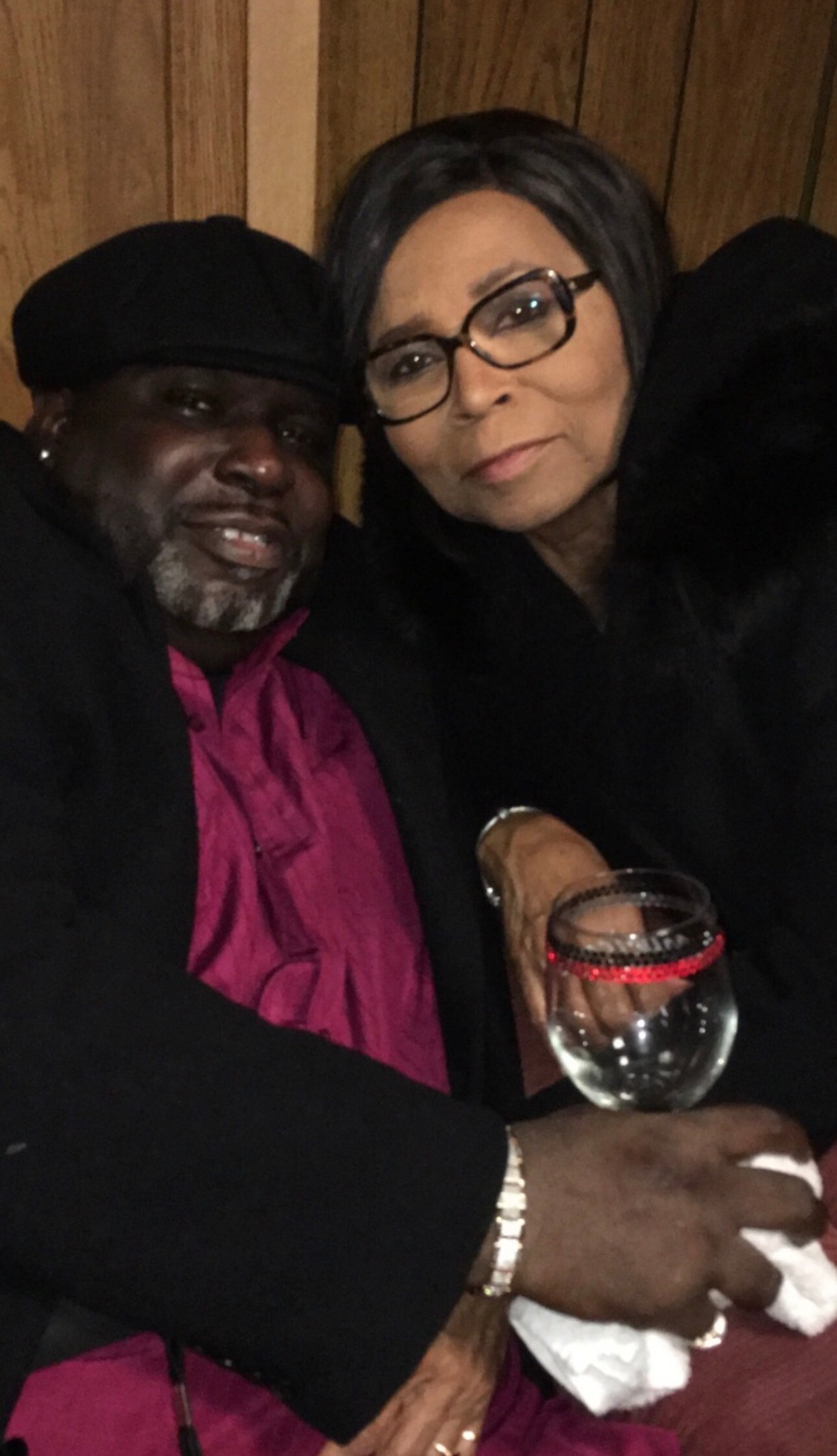 Two people posing closely for a photo, man in a hat and a woman with glasses smiling, holding a wine glass.