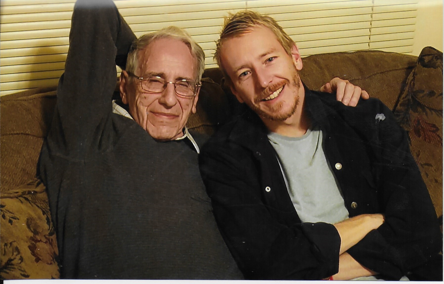Two men sitting close on a brown couch, smiling at the camera; older man with glasses on the left, younger man with blond hair on the right, arm around.
