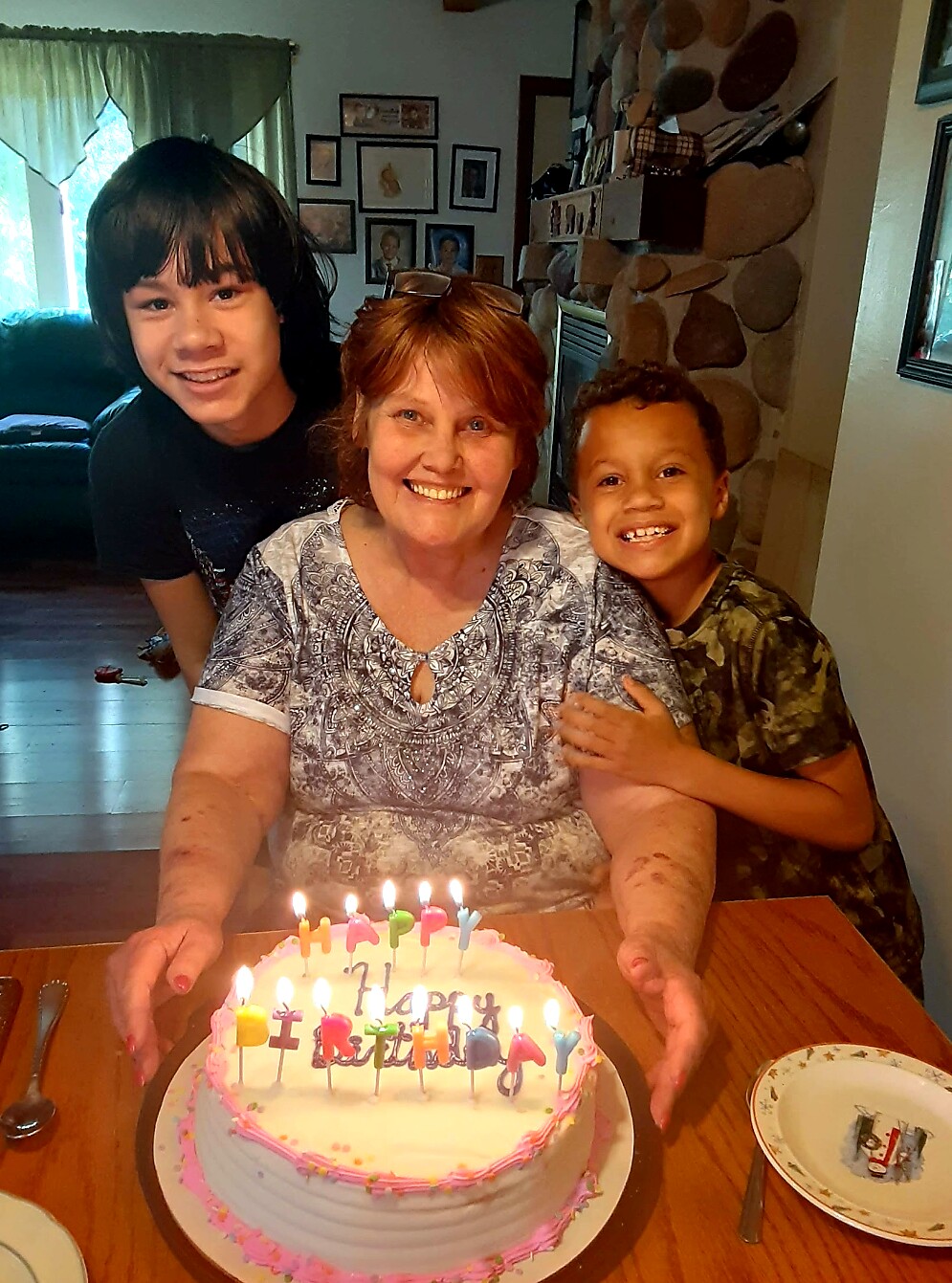 Smiling grandmother at a birthday table with two kids leaning in, in front of a pink frosted cake with candles.
