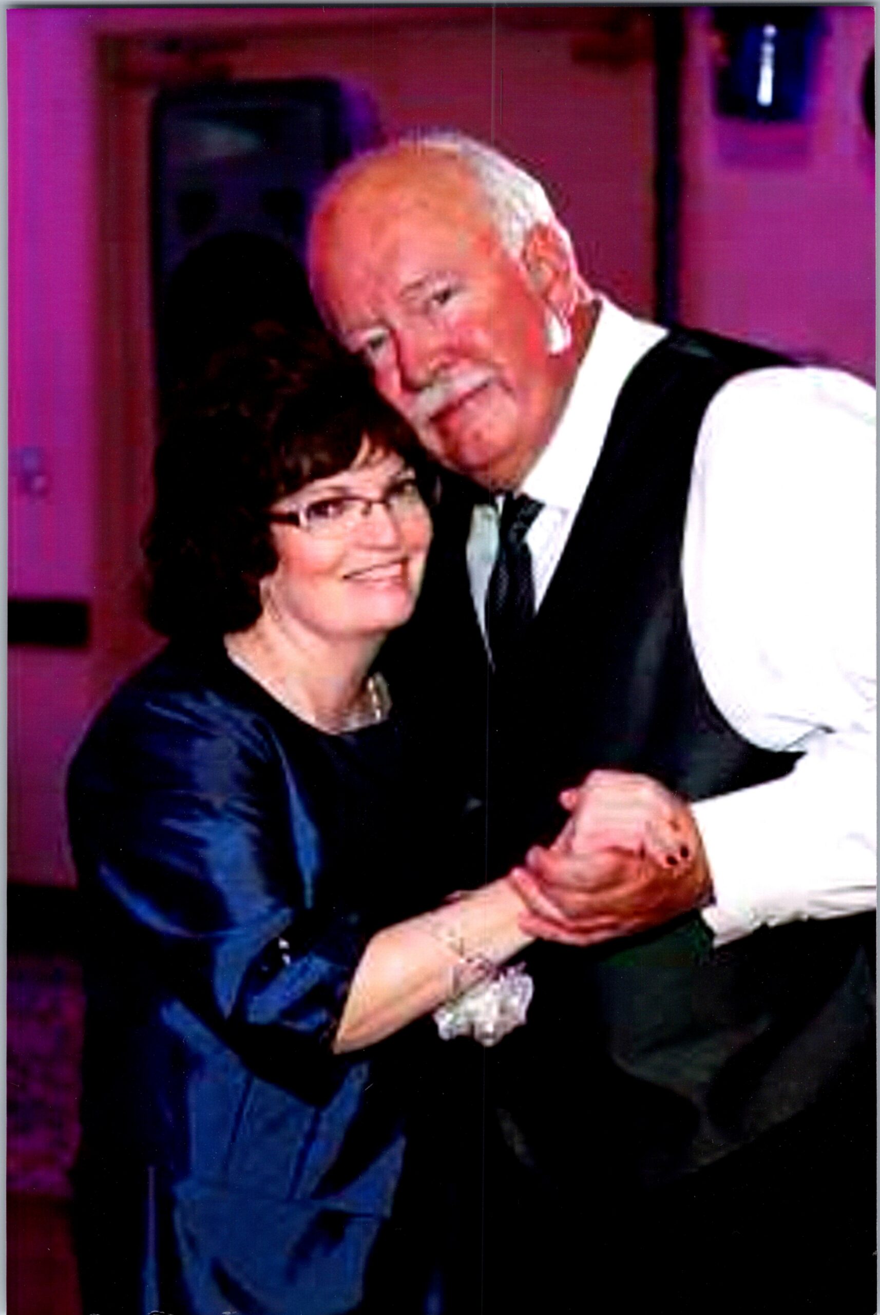 An elderly couple dancing together, smiling, dressed in formal attire with the man in a vest and the woman in a blue dress.