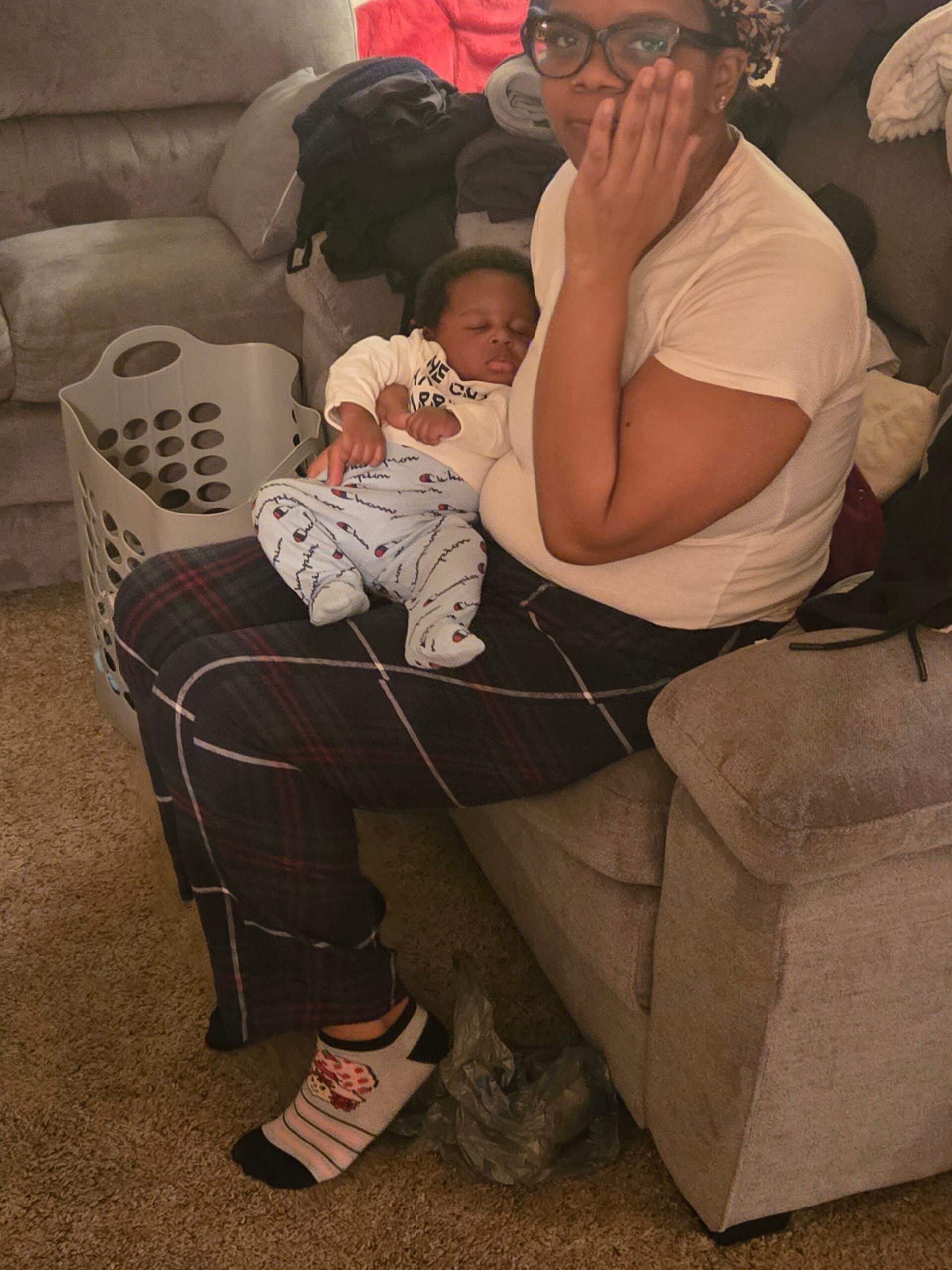 Mother wearing glasses sits on a plaid chair cradling a sleeping baby in her arms, with a laundry basket and clothes nearby in a living room.