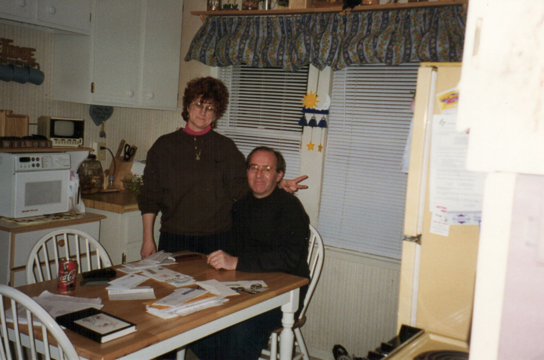 Two people, a man seated and a woman standing, pose at a wooden kitchen table scattered with papers.