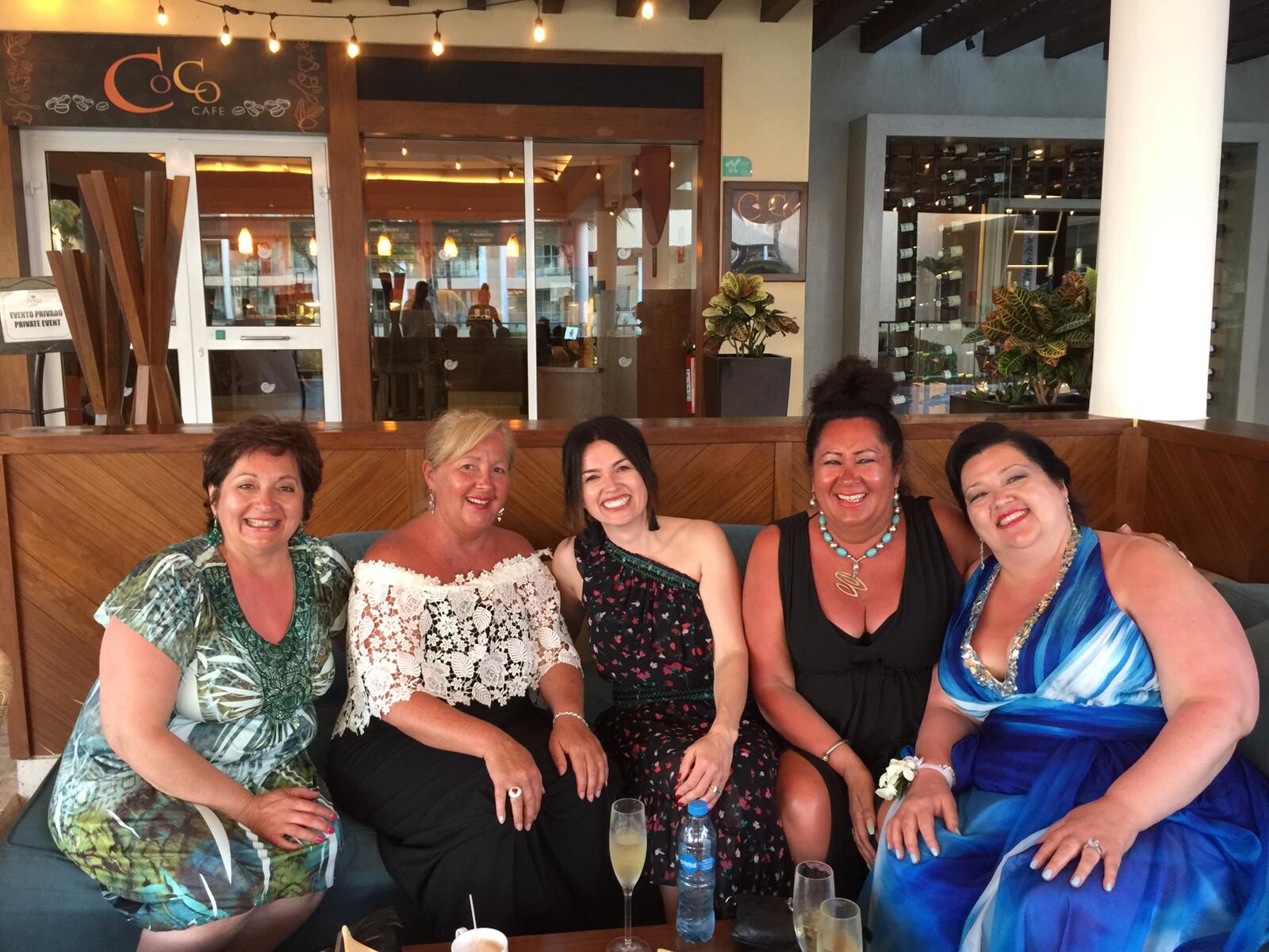 Five women in colorful dresses sit on a sofa at a cafe, smiling for the photo with drinks on the table behind them.