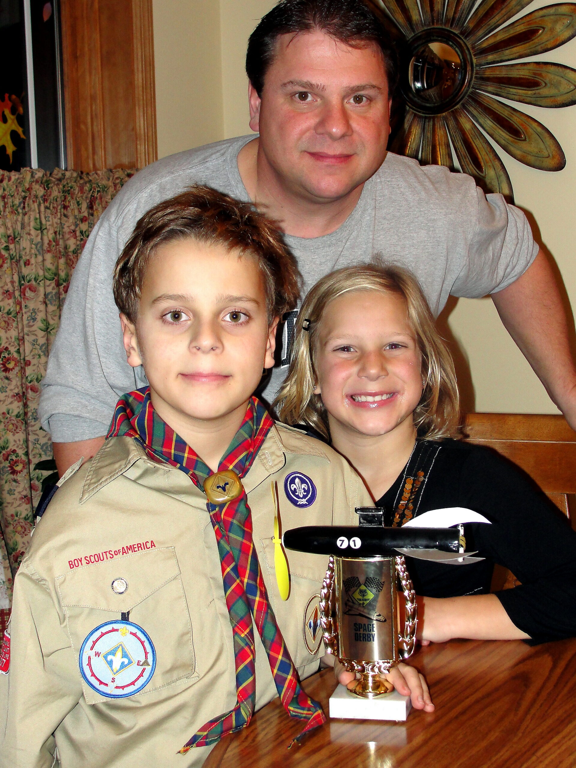 Man and two children in Boy Scouts uniforms pose with a Space Derby trophy on a wooden table.