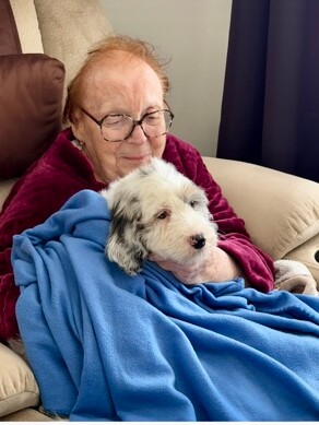 Elderly person wearing glasses sits on a beige sofa, cuddling a small white and gray dog wrapped in a blue blanket.