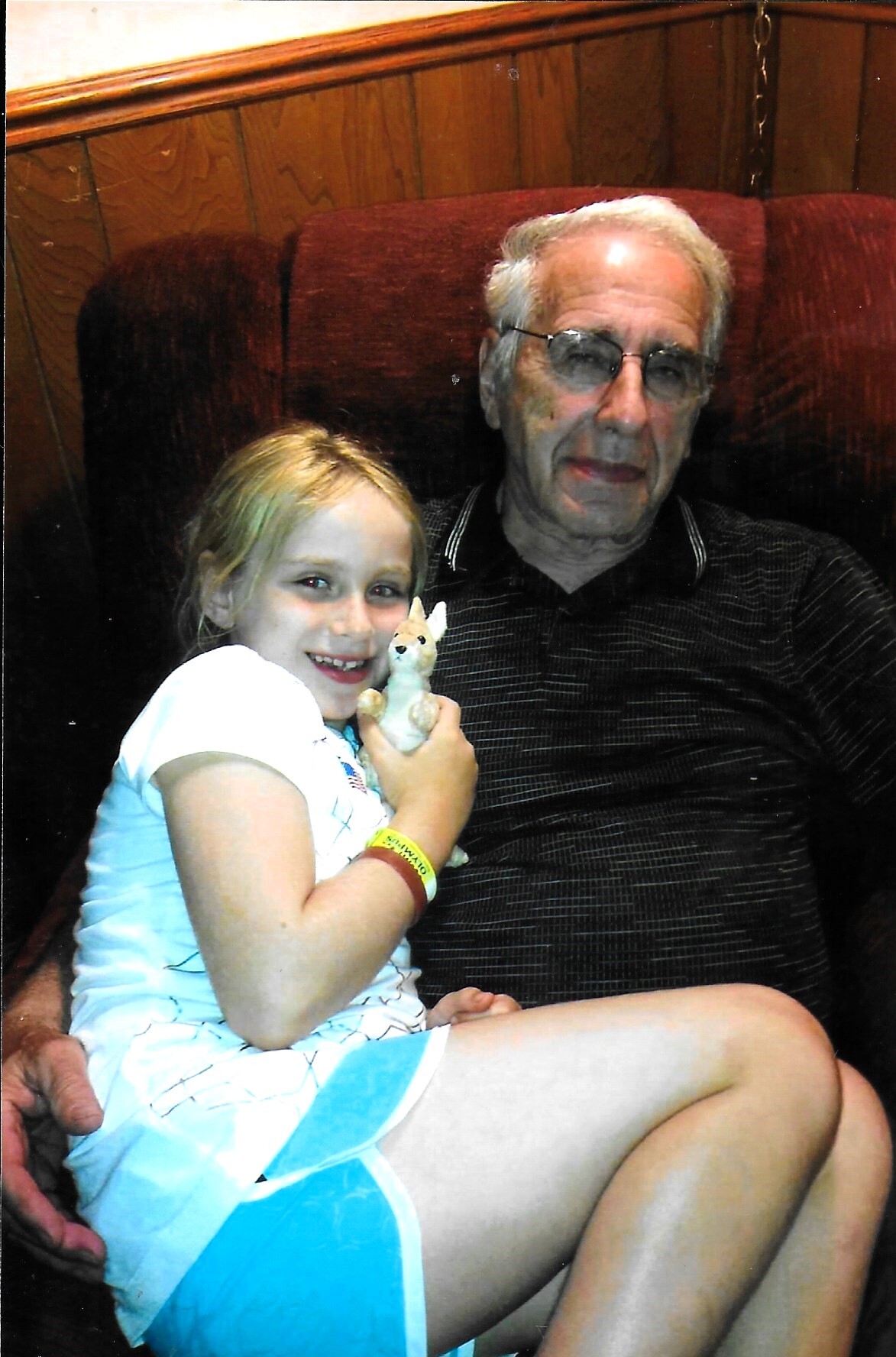 Young girl sitting on an older man's lap in a burgundy chair, holding a small plush fox toy indoors by wood-paneled walls.
