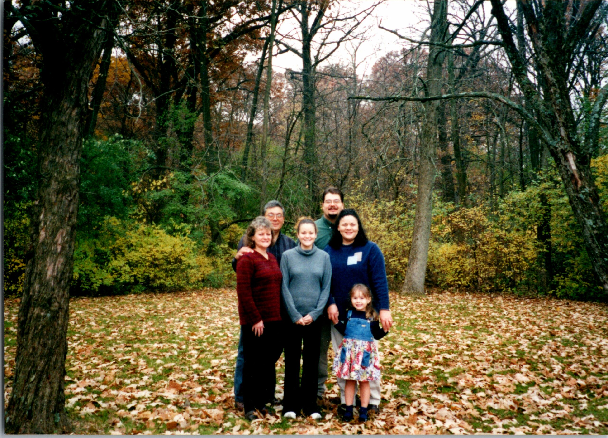 Group of six people posing together in a forest during autumn, with fallen leaves on the ground and bare trees behind them.