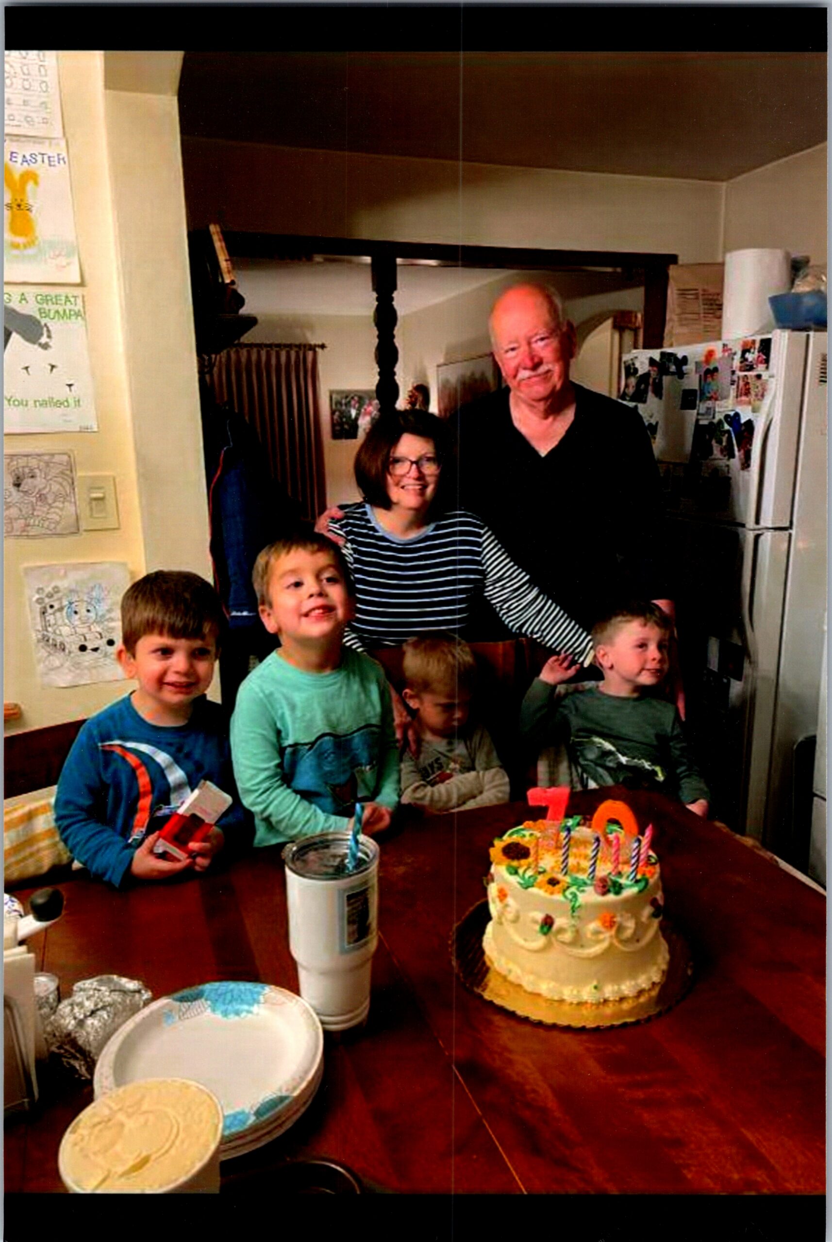 Group of children and an older couple gathered around a birthday cake with lit candles on a dining table in a kitchen.