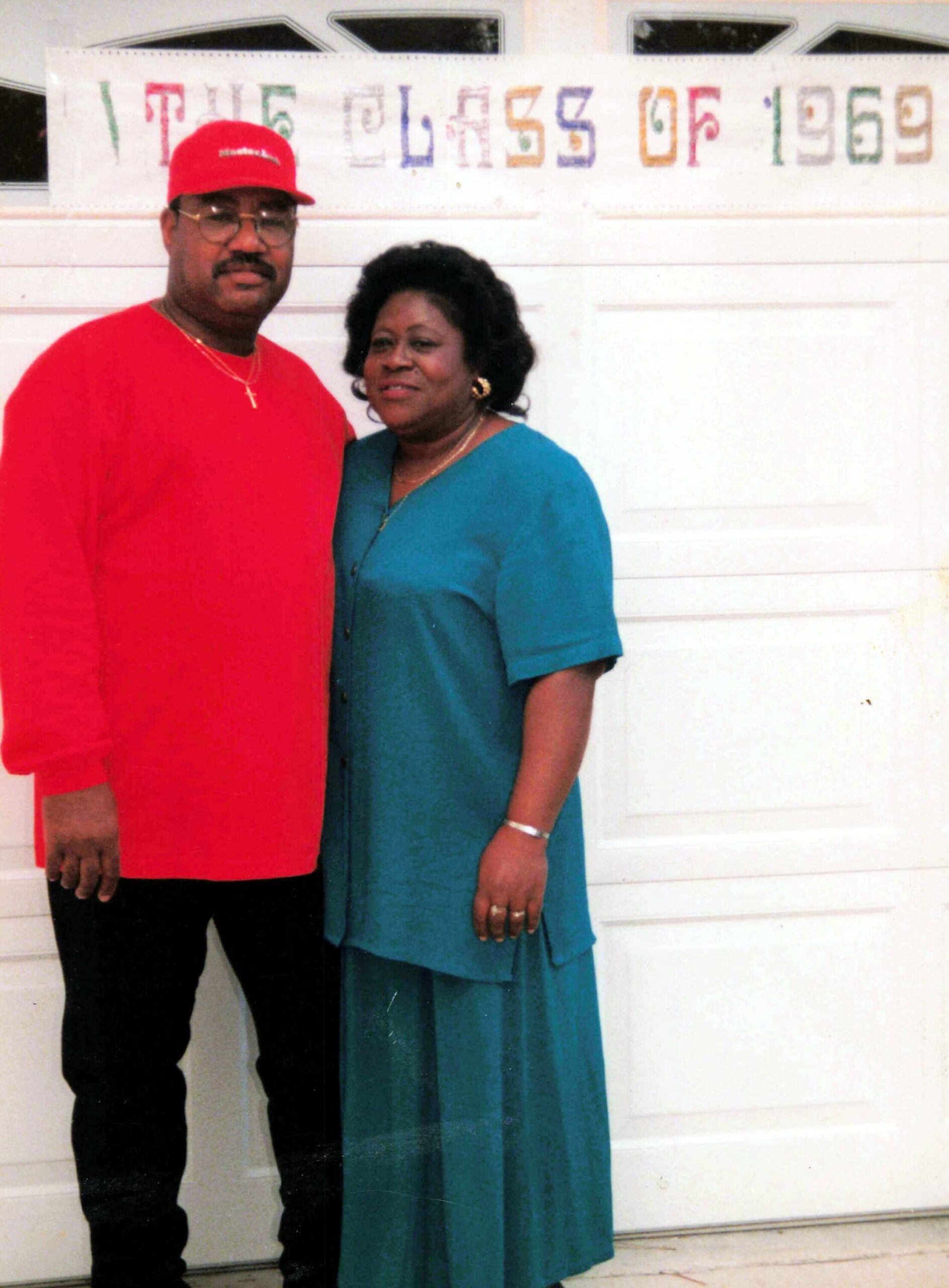 Couple posing together indoors in front of a white door; man in red shirt and cap, woman in teal dress smiling at the camera.