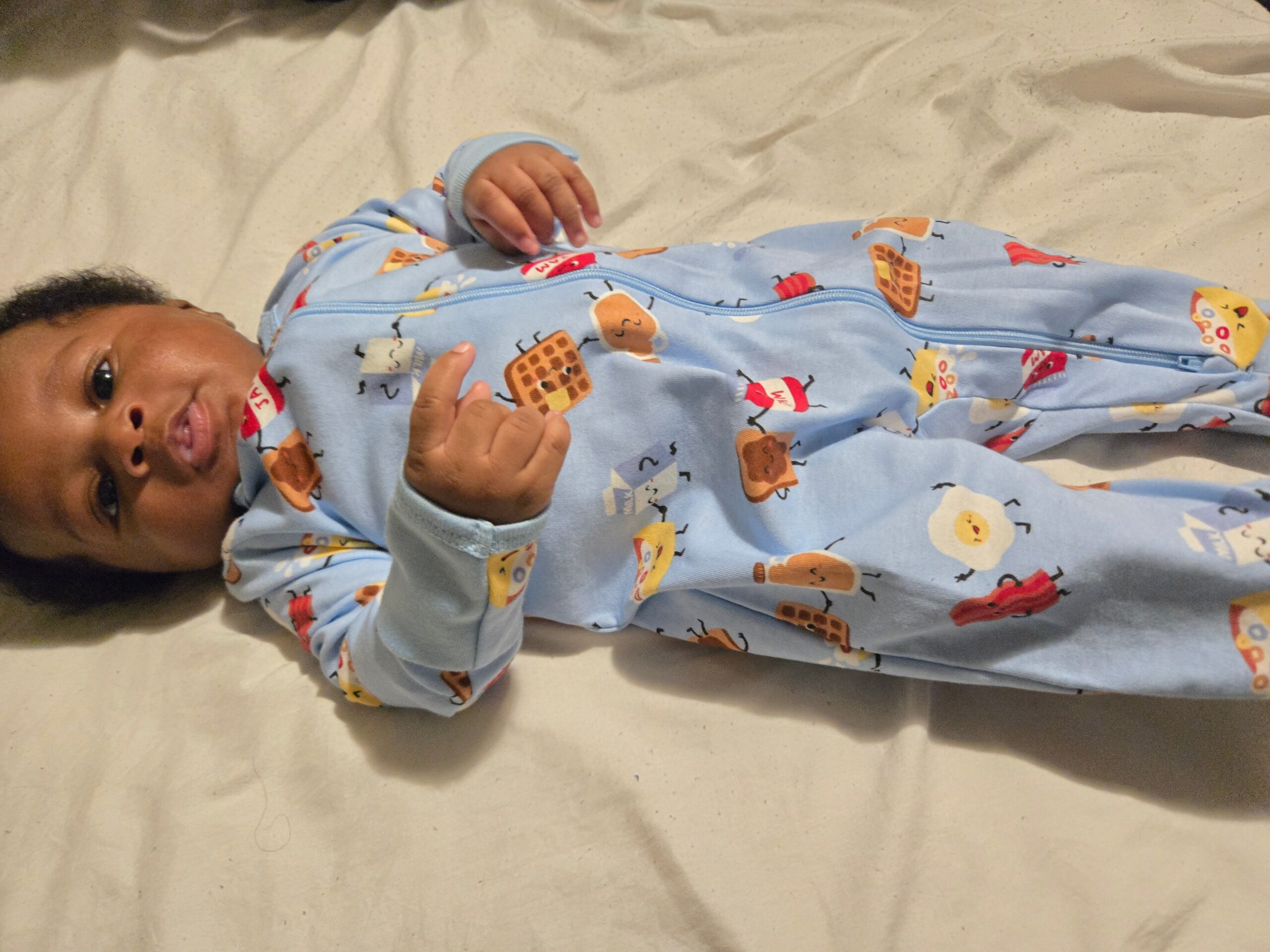 Infant lying on a pale surface in a blue sleeper with waffles and food-pattern print, looking to the side.