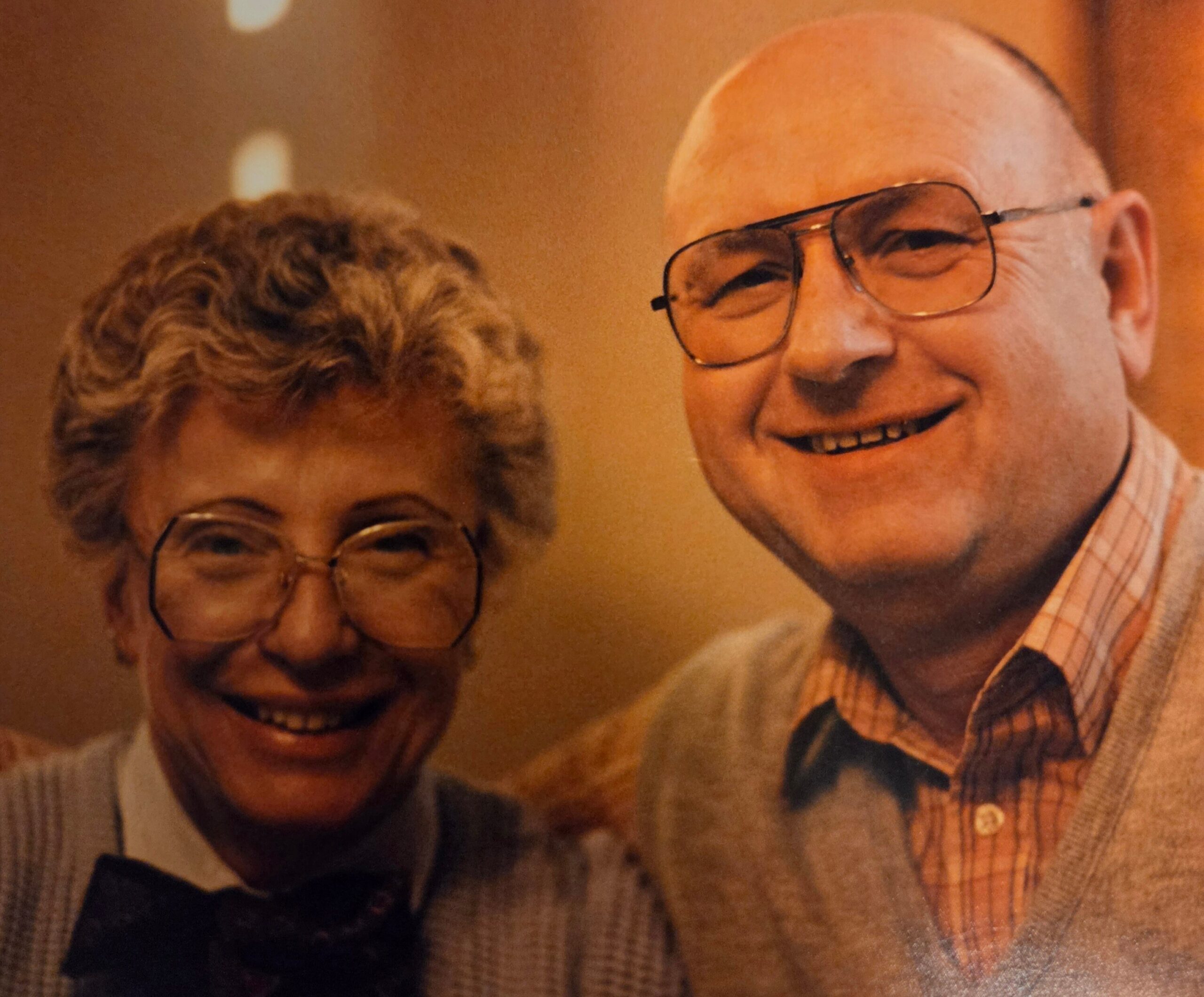 Two smiling older adults wearing glasses, close-up portrait indoors.