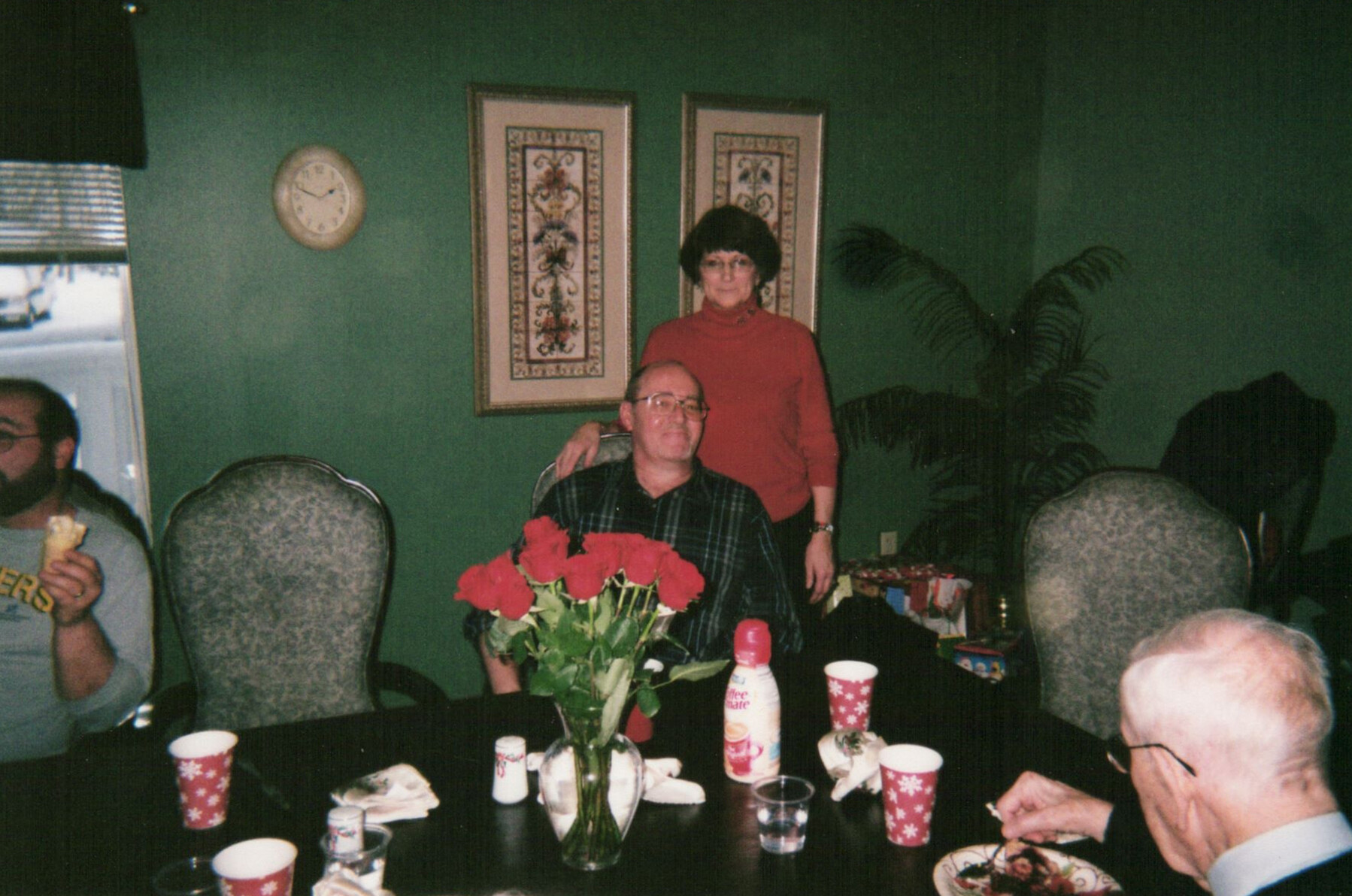 Woman in a red turtleneck stands behind a seated man at a family dinner around a table with a vase of red roses.