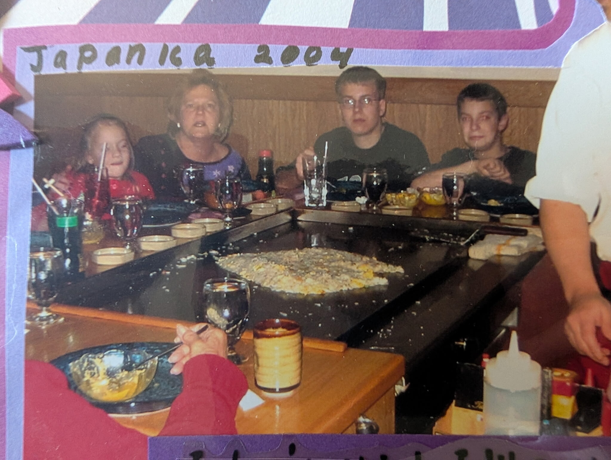 Family of four sits at a banquet-style table with plates and glasses, a girl on the left, adults in the center and right, food and drinks in front.