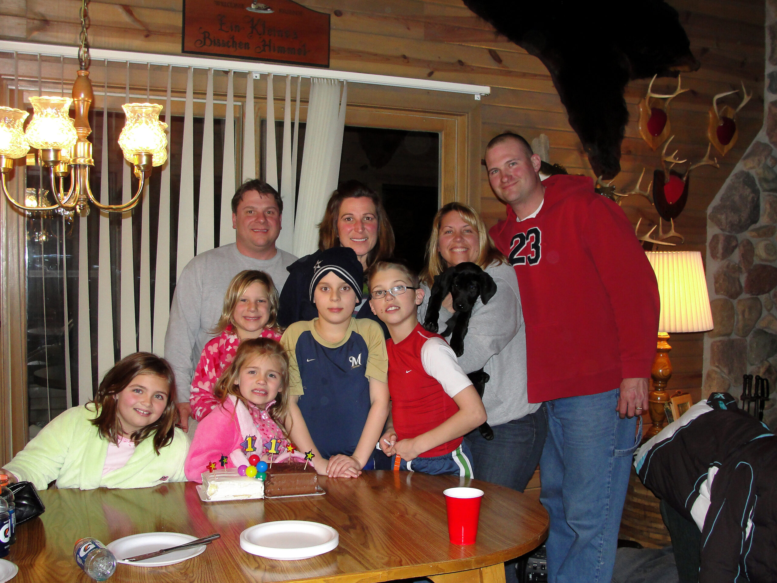 Family and friends pose around a cake on a wooden table at a cabin birthday party.