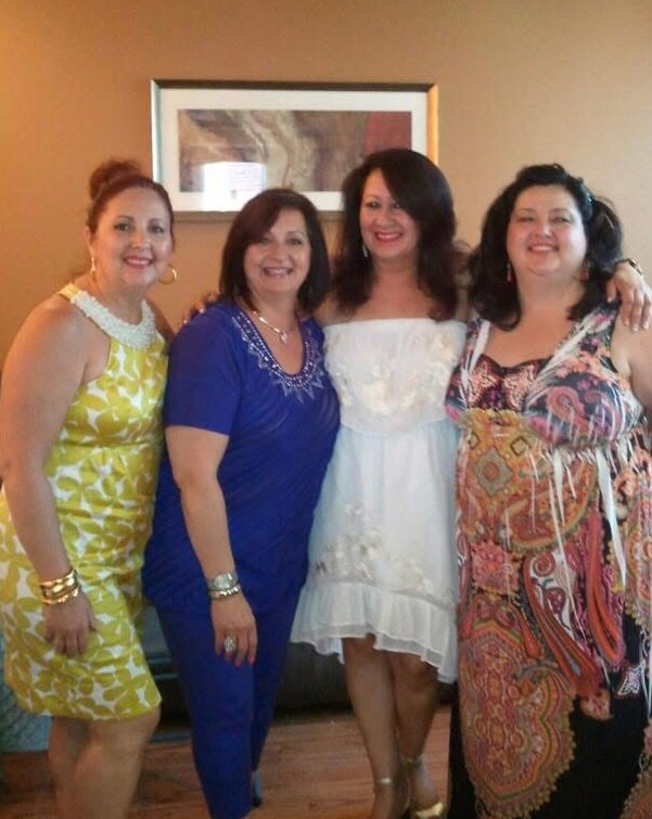 Four women stand side by side indoors, smiling for a group photo; one in a yellow dress, one in blue, one in a white dress, and another in a colorful patterned dress with jewelry.