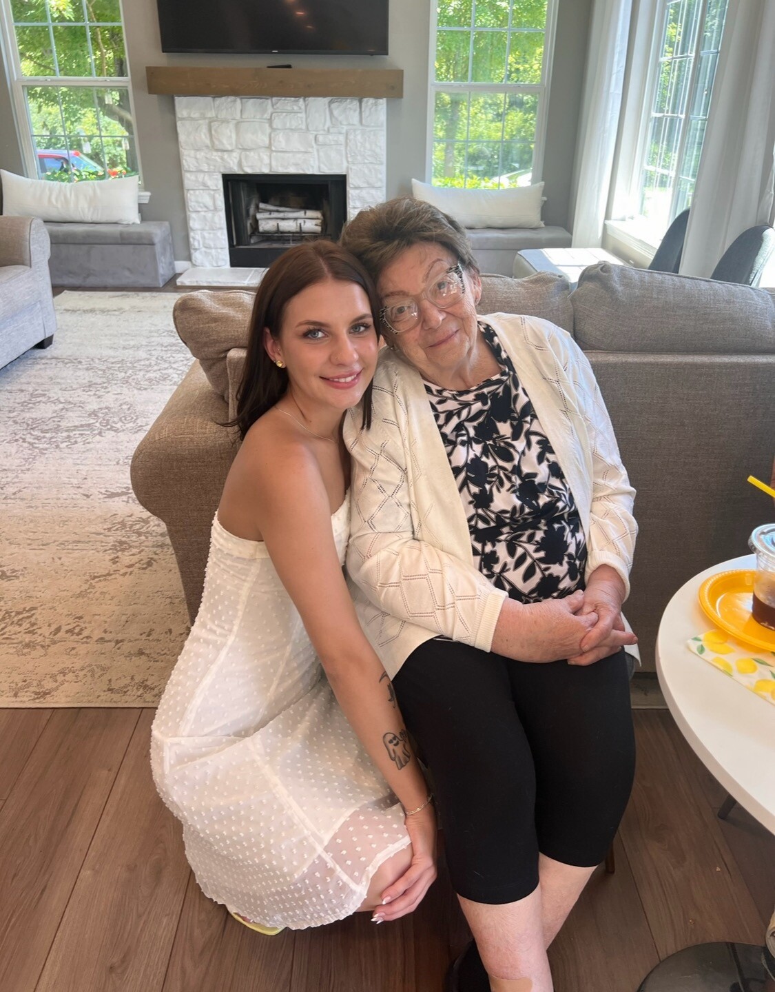 Young woman in a white dress sits beside an elderly woman on a sofa, smiling together in a bright living room with a stone fireplace and large windows behind them.