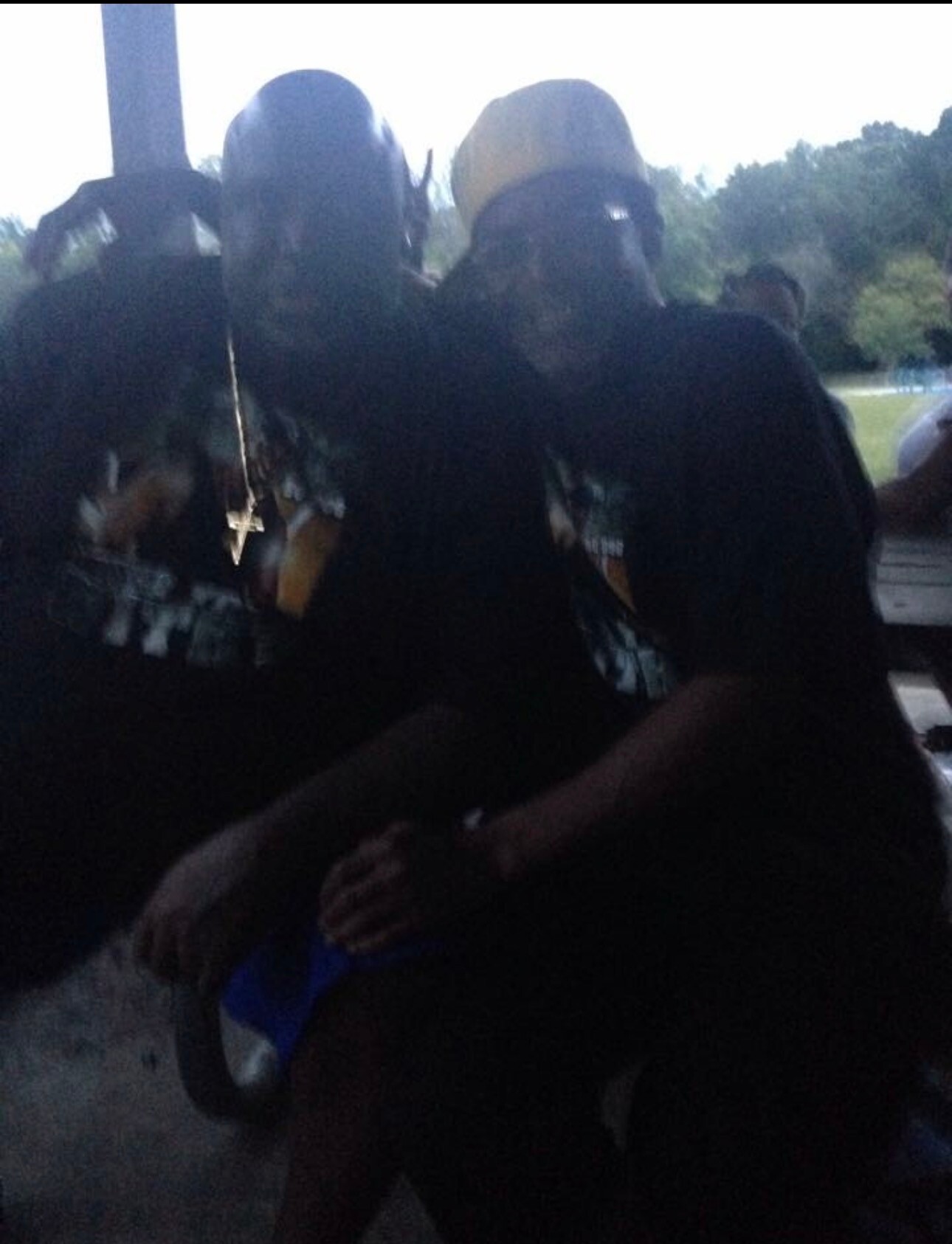 Two people in dark clothing wearing full-face gas masks pose for a photo on a shaded porch outdoors.