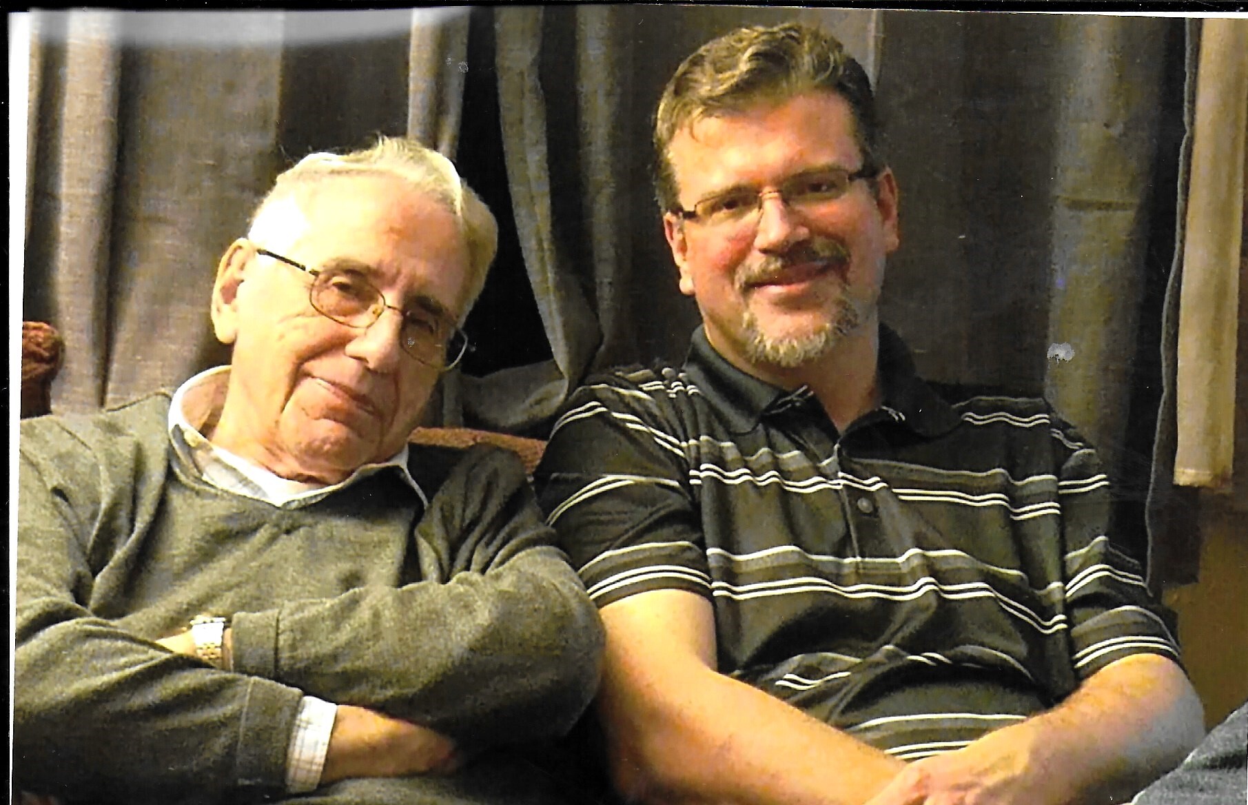 Two men sit closely on a couch, smiling at the camera; older man on the left with glasses and gray hair, younger man on the right with a striped polo shirt and beard.