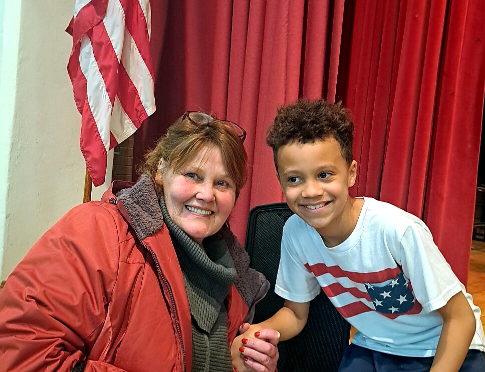 Smiling woman in a red jacket sits beside a boy in a white shirt with a USA flag graphic; red curtains backdrop and flag visible behind them.