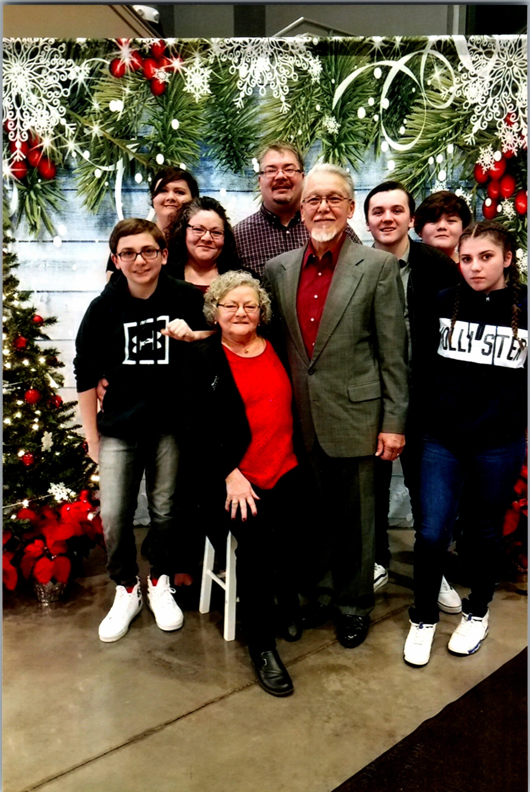 Group of people posing together in front of a festive Christmas backdrop with greenery and snowflake decor while a decorated tree stands nearby.