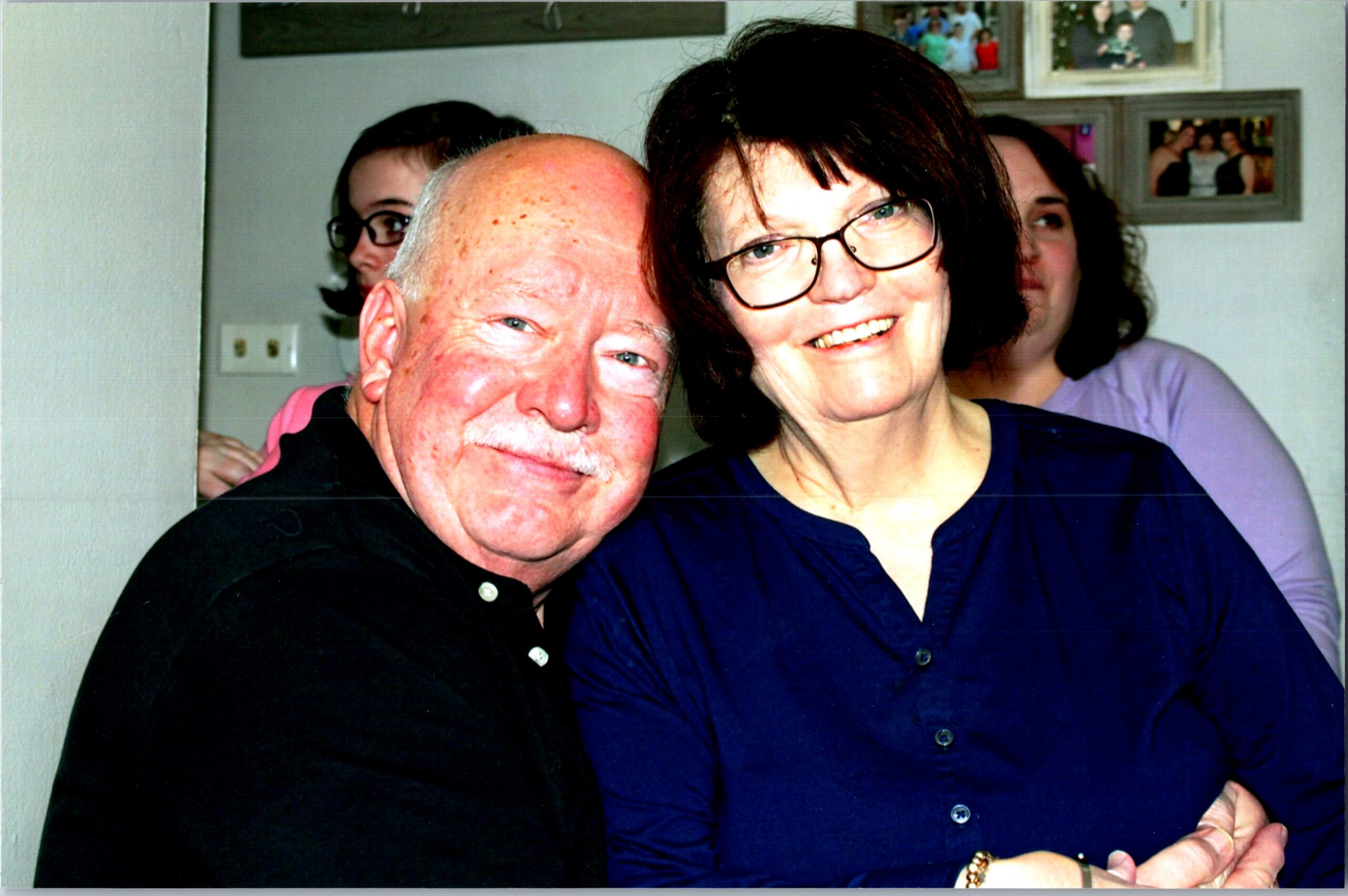 Smiling older man and woman posing closely for a photo indoors, with family photos on the wall behind them.