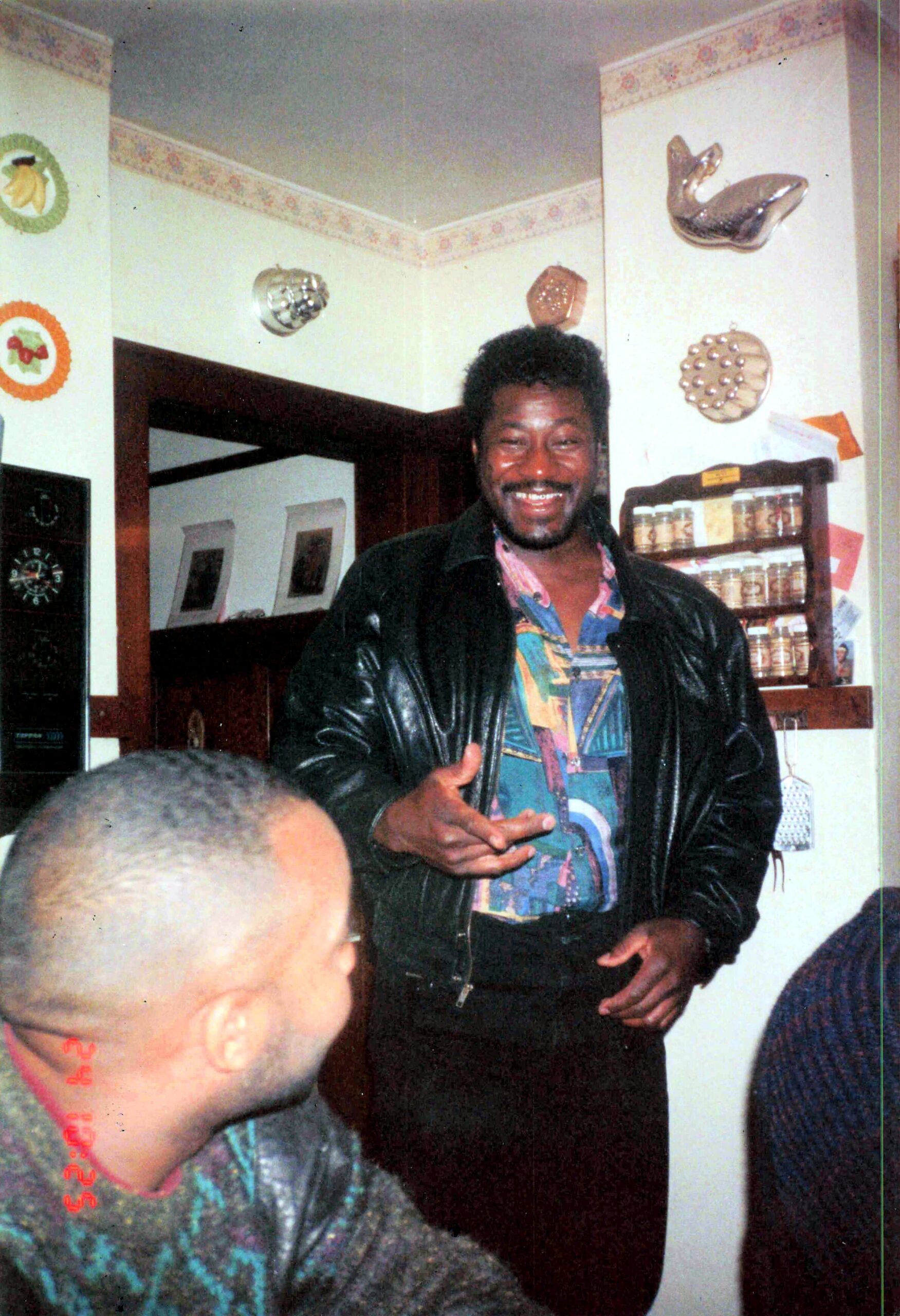 Smiling man in a colorful shirt and black jacket poses in a cozy kitchen with decorative plates on the wall; others are partially visible in the foreground.
