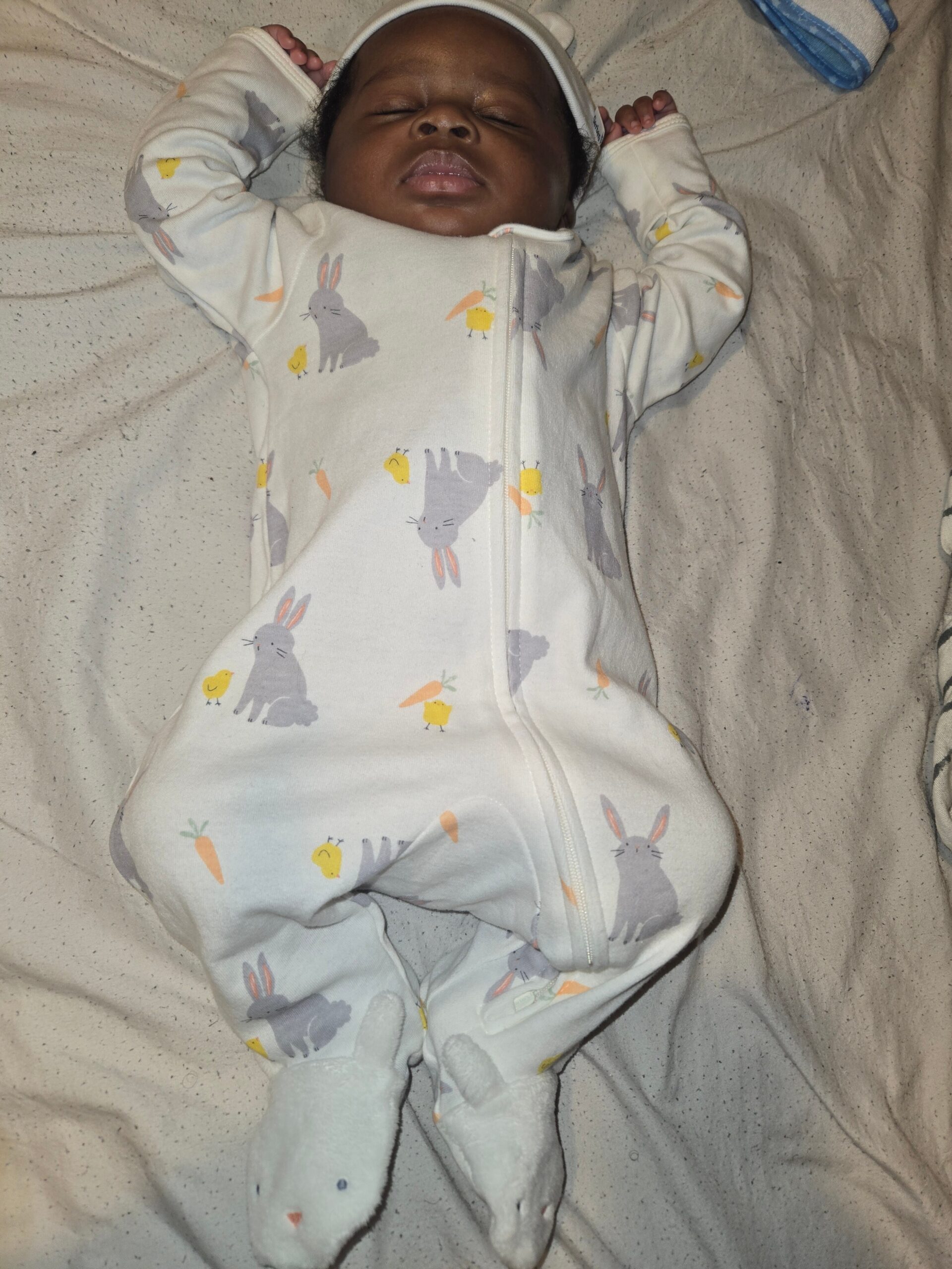 Sleeping baby in a white bunny-print onesie with arms stretched above head on a bed