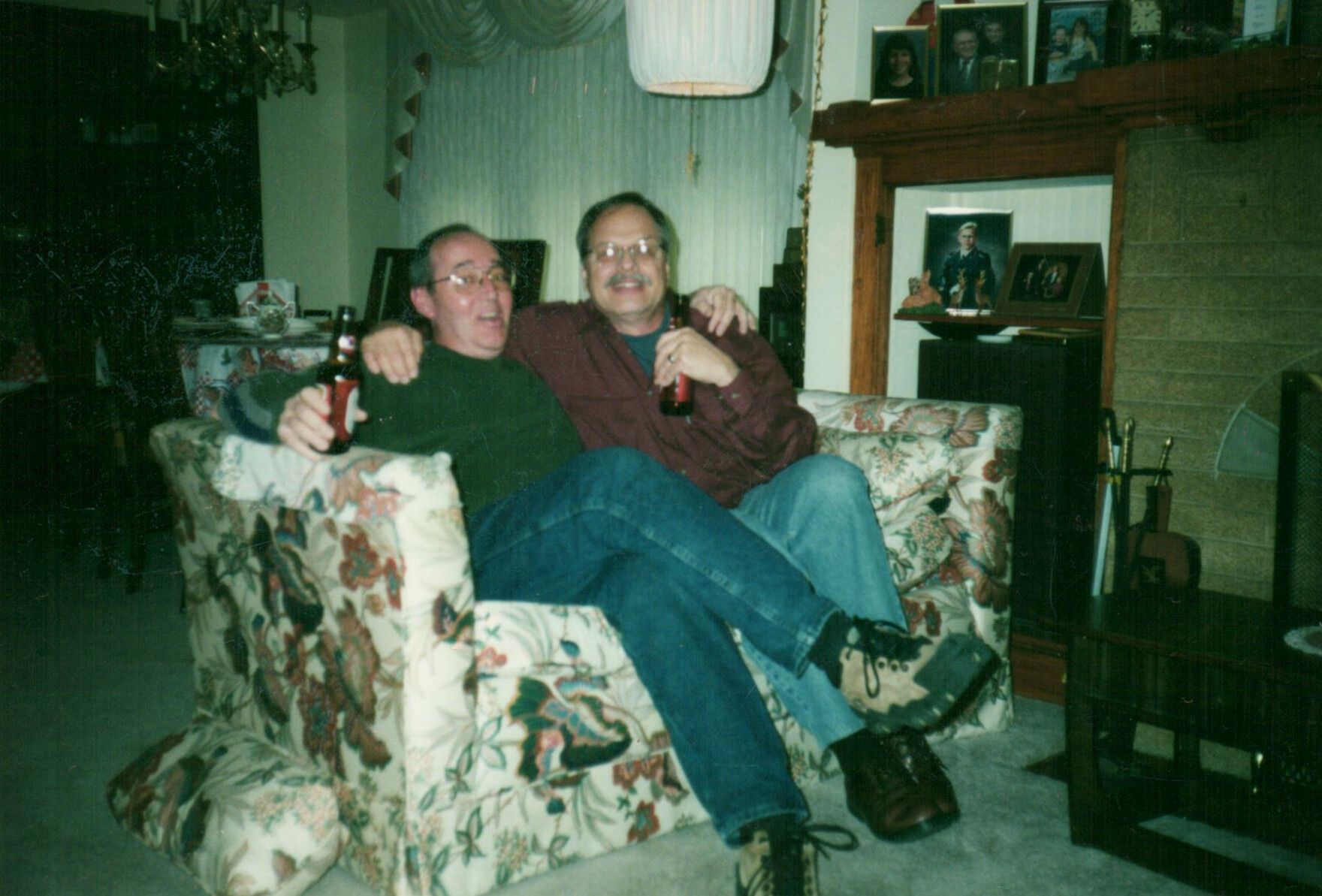 Two men sit close on a floral sofa, each holding a beer and smiling at the camera in a cozy living room setting.