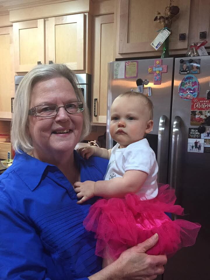 Smiling grandmother in a blue blouse holds a baby in a pink tutu in a kitchen near a stainless fridge with magnets and notes.