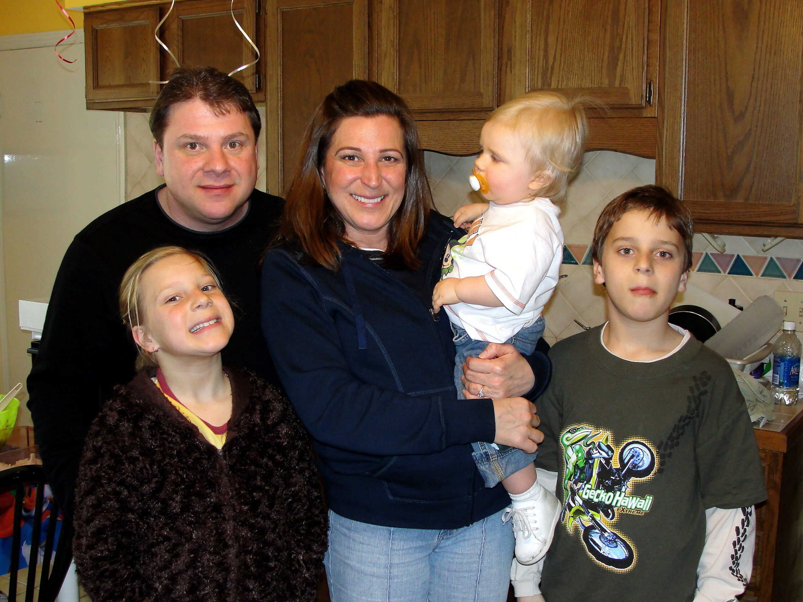 Family of five posing together in a kitchen, smiling at the camera; a man and woman hold a toddler, with two older children nearby.