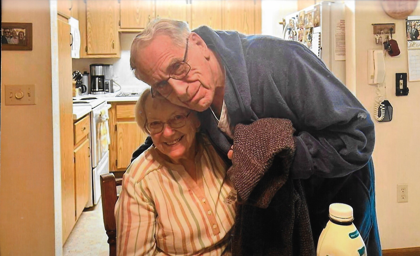 An elderly couple smiles in a cozy kitchen; the man leans over the seated woman wrapped in a robe, holding a sweater and a bottle on the table nearby.