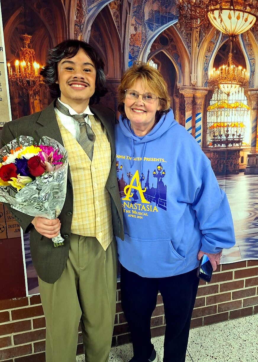 Young man in a 1920s-style suit holds a bouquet and poses with a smiling woman in a blue Anastasia hoodie, in front of an ornate backdrop.