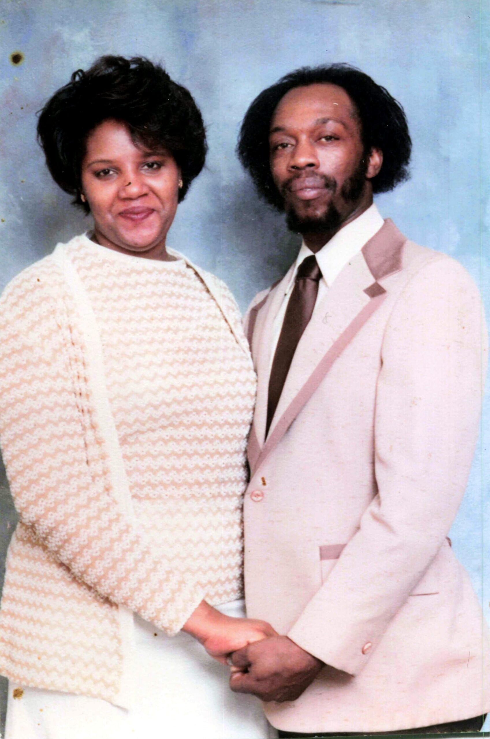 Smiling couple in 1970s attire posing together against a blue backdrop, woman in a cream knit cardigan and man in a pale pink suit.